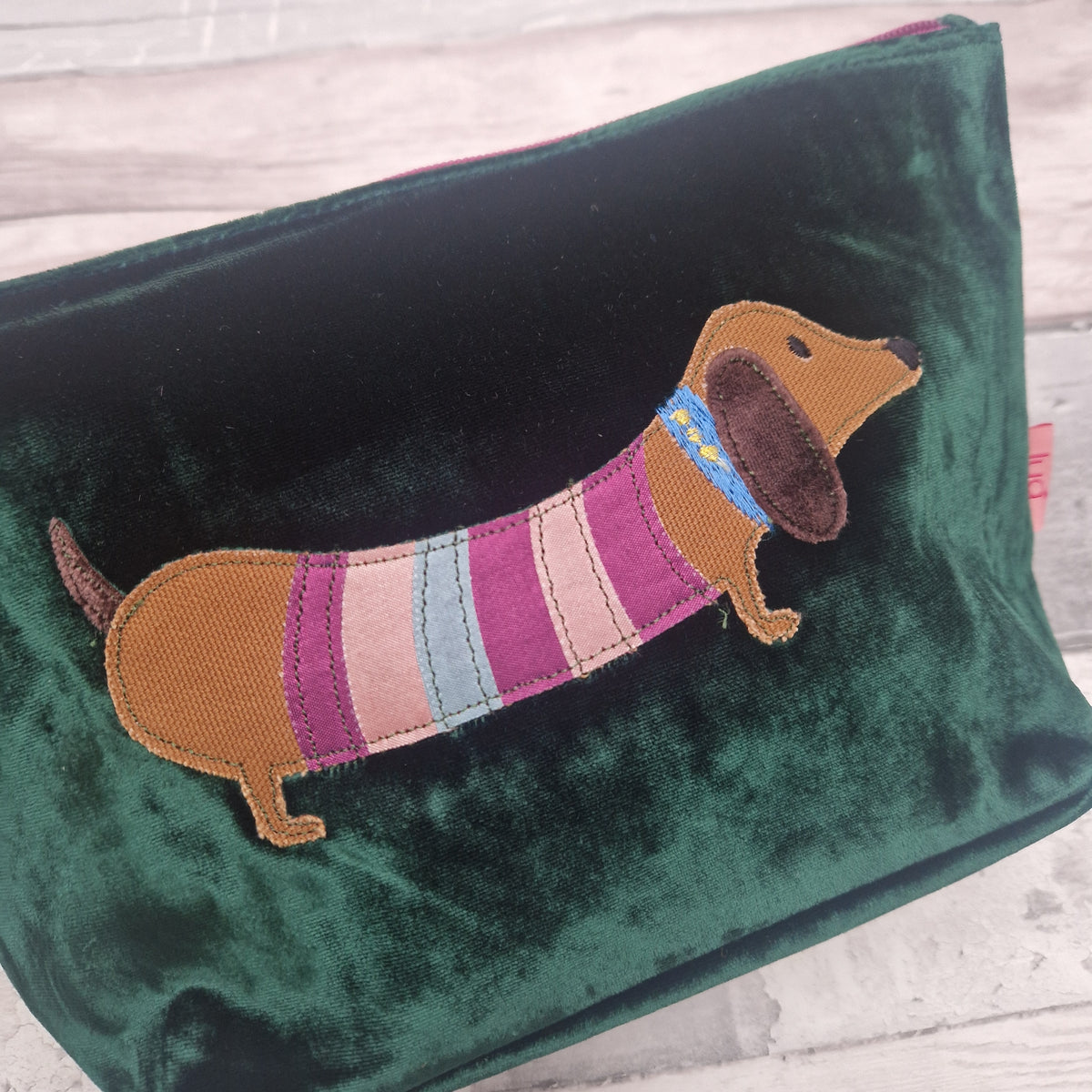 A dark green velvet cosmetic pouch featuring an embroidered dachshund dog wearing a pink, purple, and blue striped sweater, set against a light wood background.