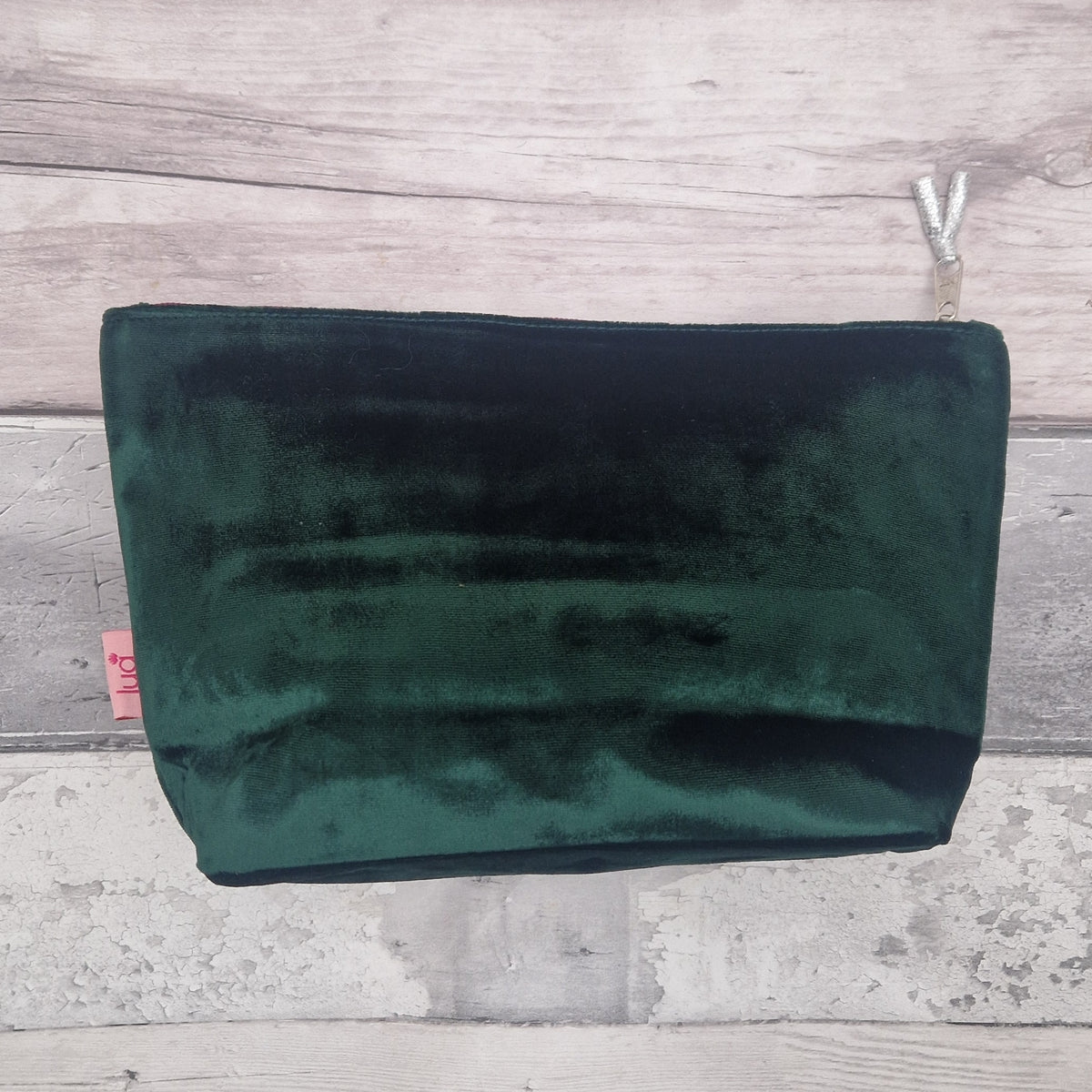 A dark forest green velvet pouch or clutch with a silver zipper pull, displayed flat against a light gray wood plank background.