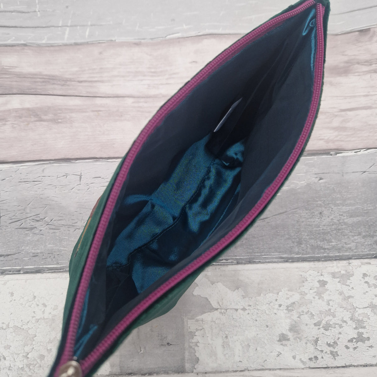 An open green pouch or clutch with a dark teal interior lining and a pink zipper, shown from above on a light gray wood-plank surface.