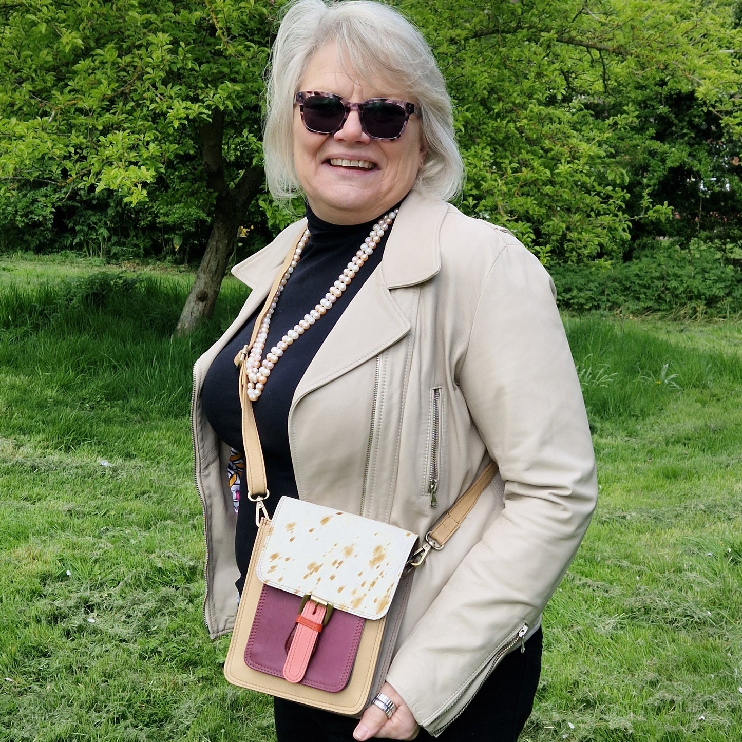 Woman standing outdoors wearing sunglasses and a beige jacket with a colorful handbag.