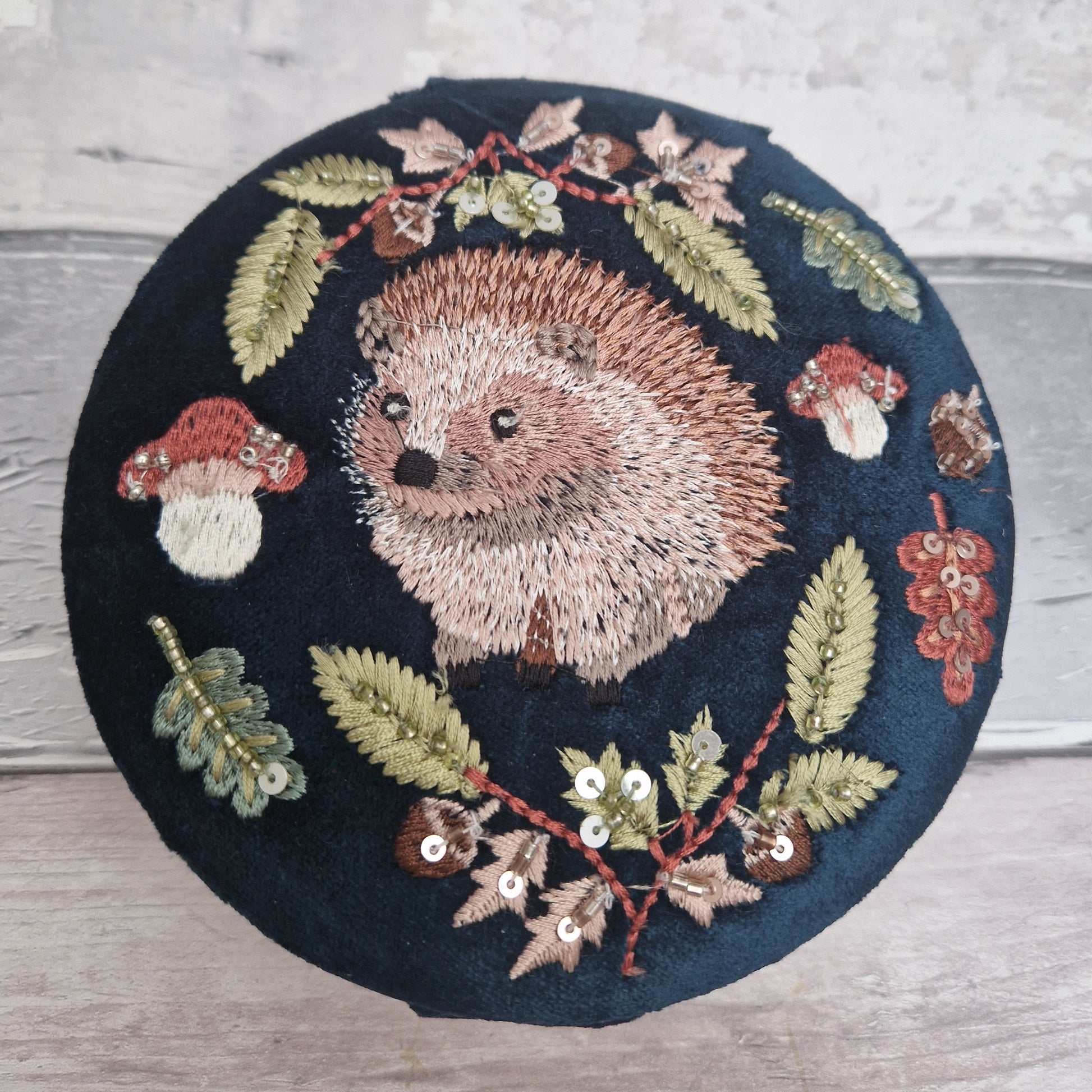 Navy velvet covered jewellery bow with embroidered scene of a Hedgehog amongst woodland leaves and mushrooms. The design is finished with sequins and beads.