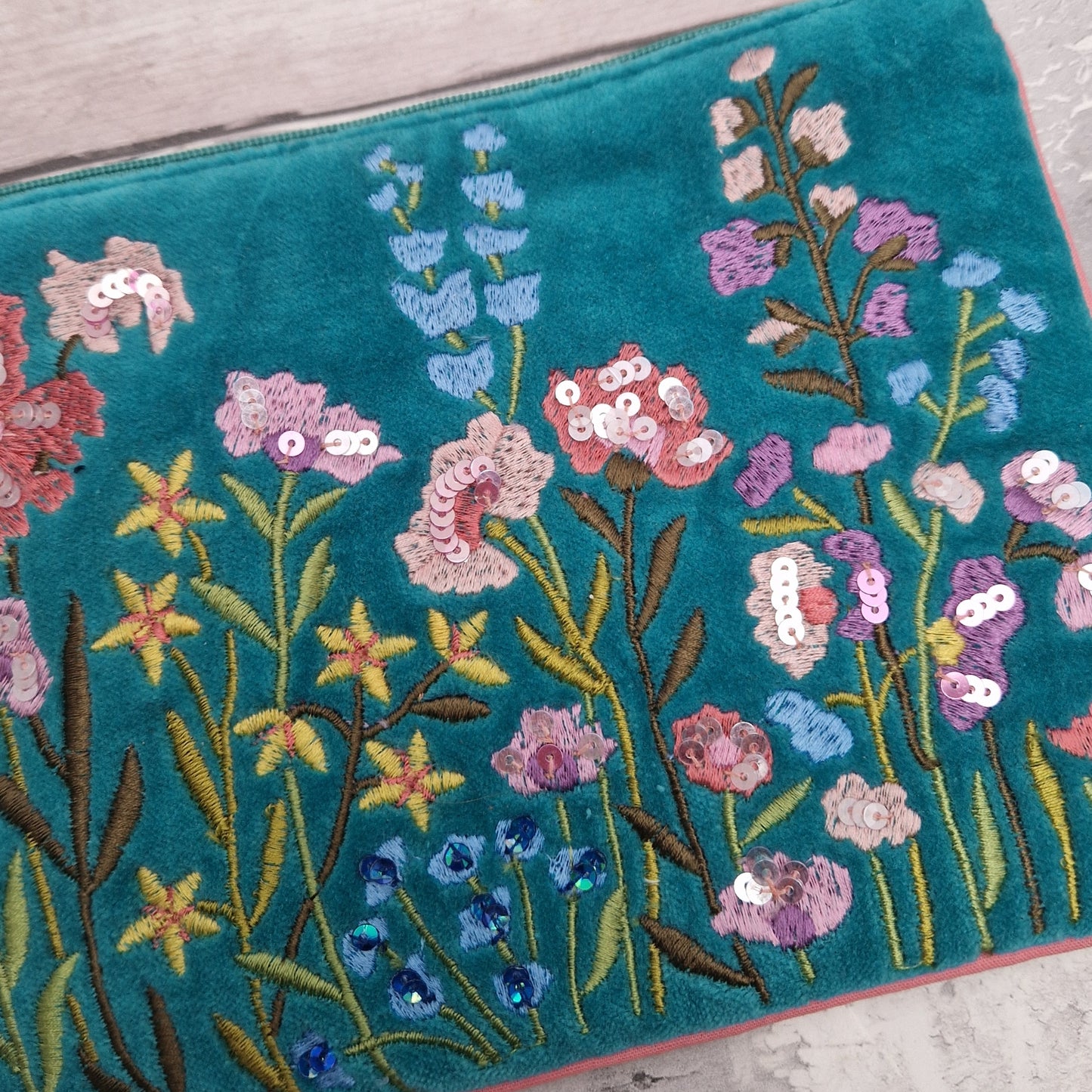 Turquoise velvet pouch decorated with embroidered flowers in pink and yellow. The pouch is finished in pink piping, a pink tassel and metallic sequins.