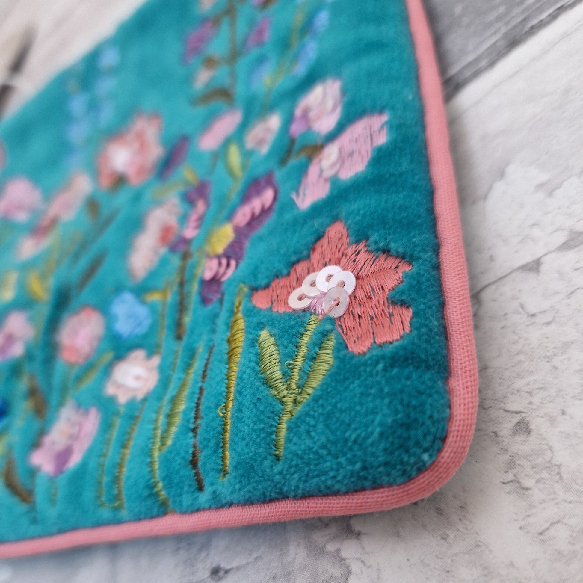 Turquoise velvet pouch decorated with embroidered flowers in pink and yellow. The pouch is finished in pink piping, a pink tassel and metallic sequins.