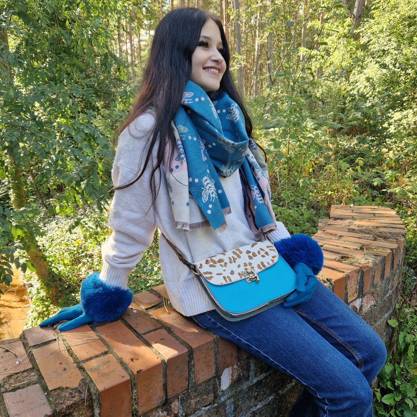 Woman sitting on a brick wall outdoors wearing a blue scarf, gloves, and a blue handbag.