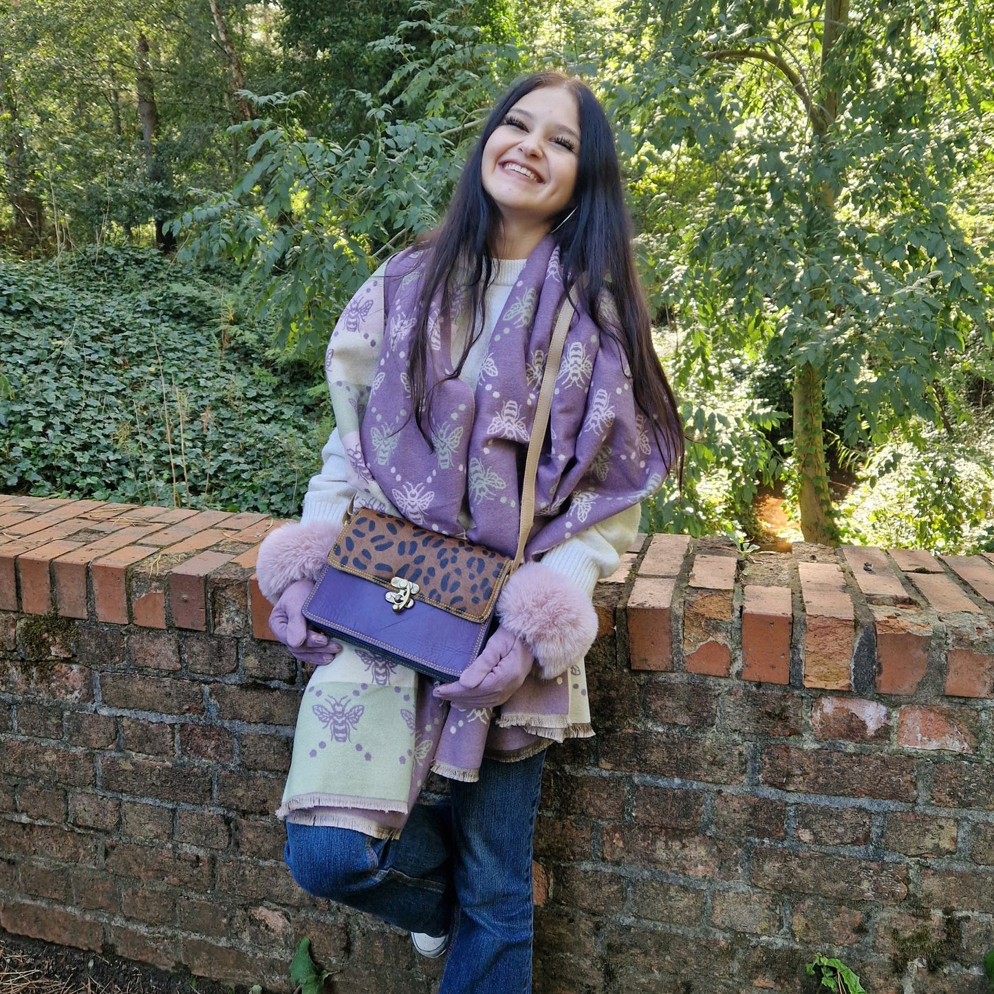 Woman wearing a purple scarf and holding a matching bag against a brick wall with greenery.