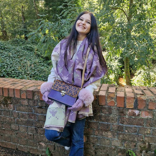 Woman wearing a purple scarf and holding a matching bag against a brick wall with greenery.