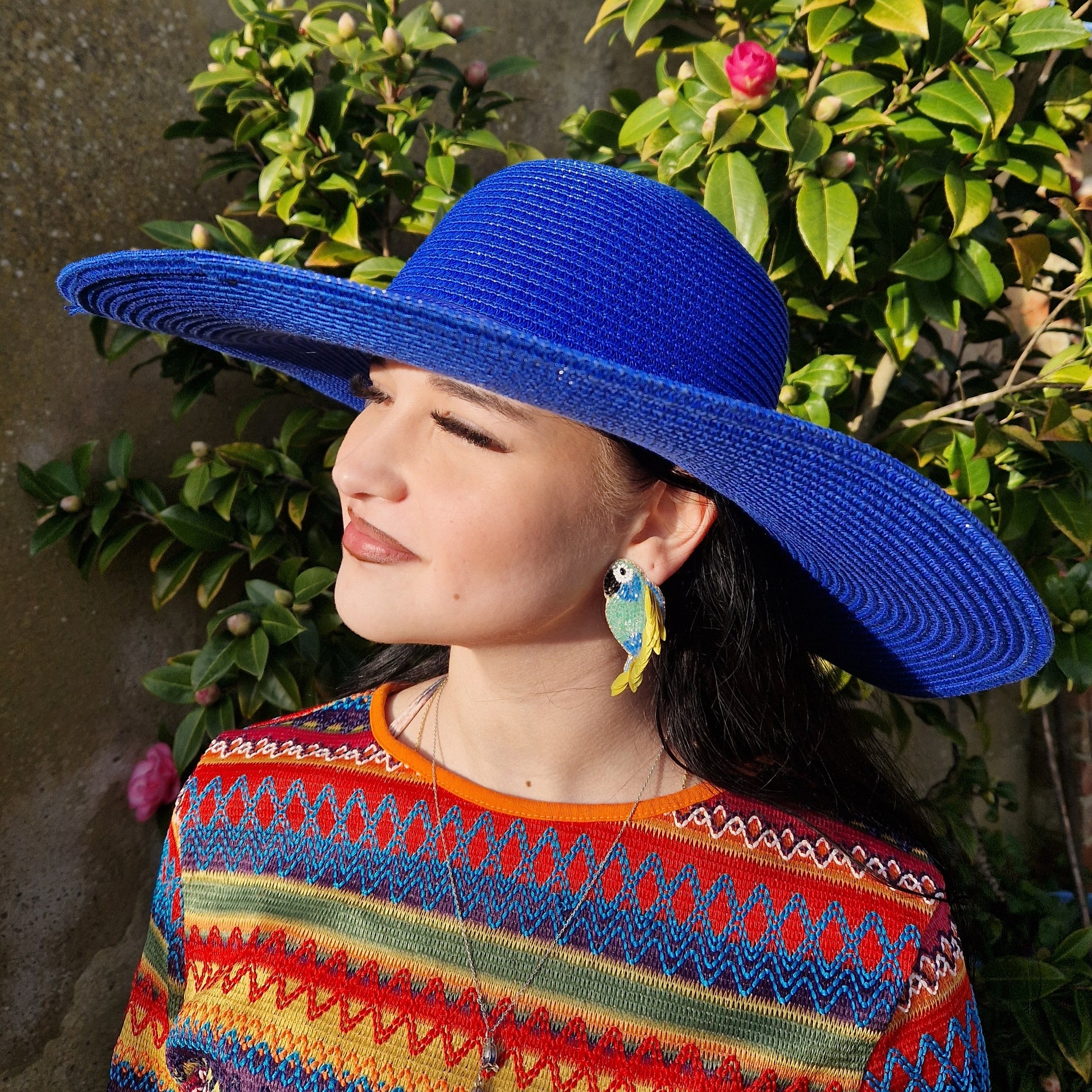 Young woman wearing a wide brimmed electric blue sun hat and beaded parrot shaped earrings in blue and green.