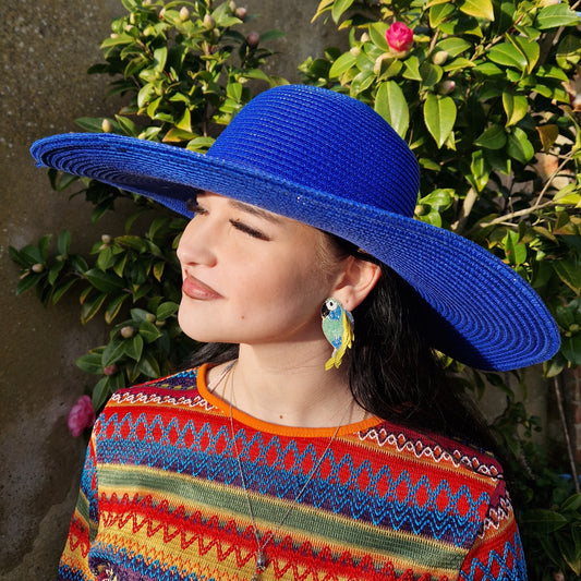 Young woman wearing a wide brimmed electric blue sun hat and beaded parrot shaped earrings in blue and green.