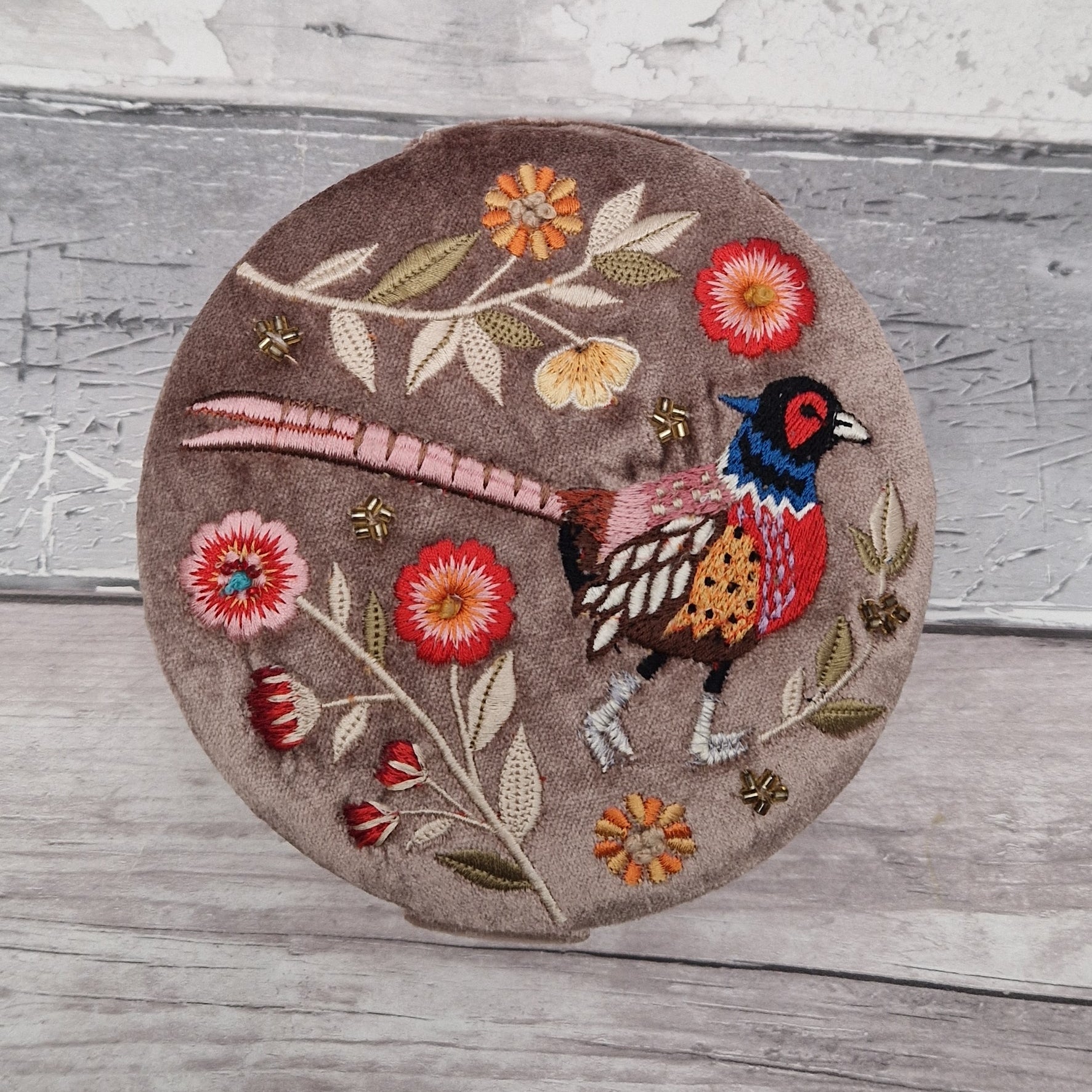 Round velvet jewellery box in a fawn colour. Decorated with an embroidered scene of a pheasant and flowers. Finished with golden beads.