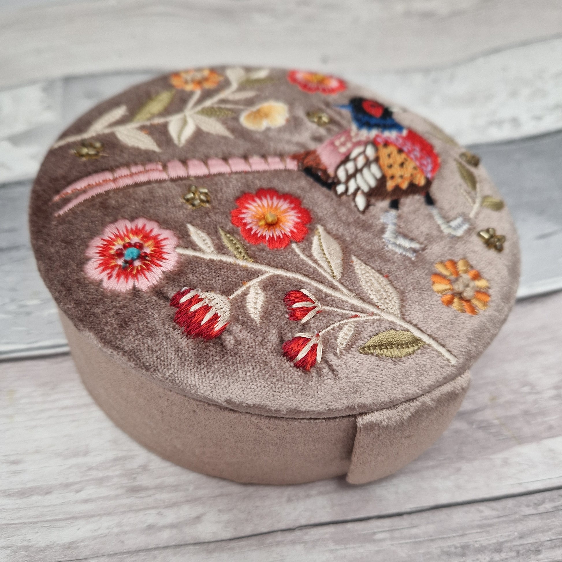 Round velvet jewellery box in a fawn colour. Decorated with an embroidered scene of a pheasant and flowers. Finished with golden beads.