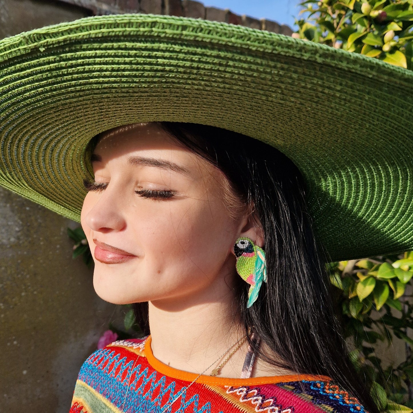 Young woman wearing a wide brimmed emerald green sun hat and beaded parrot shaped earrings in pink and green.