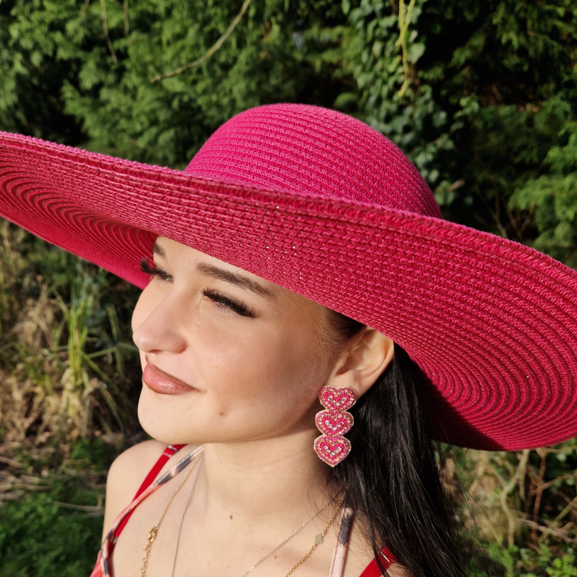 Young woman wearing a wide brimmed hot pink sunhat and pink beaded love heart earrings.