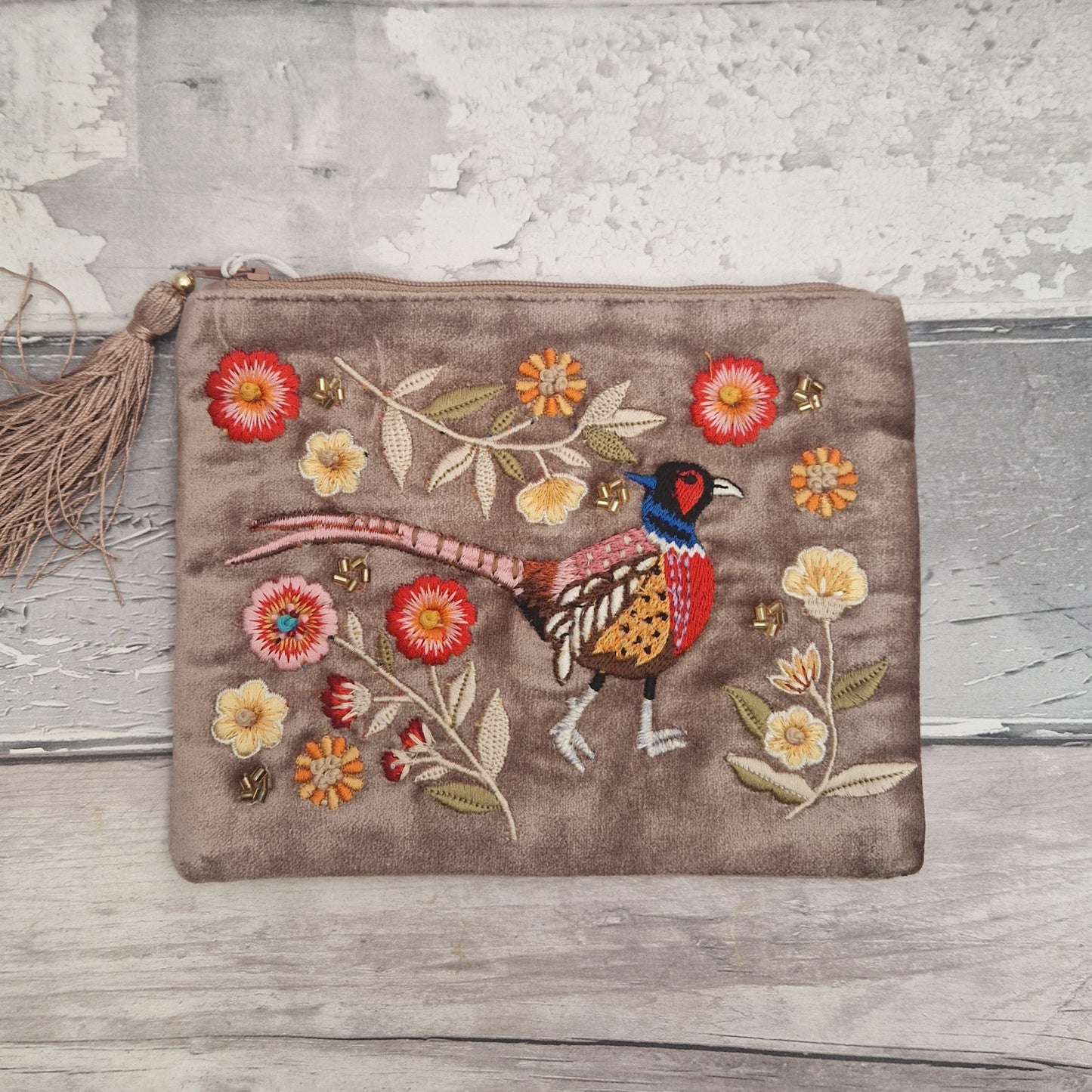 A rectangular taupe velvet pouch with a tassel zipper pull, featuring colorful embroidered pheasant and flowers on the front, set against a rustic distressed wood background.