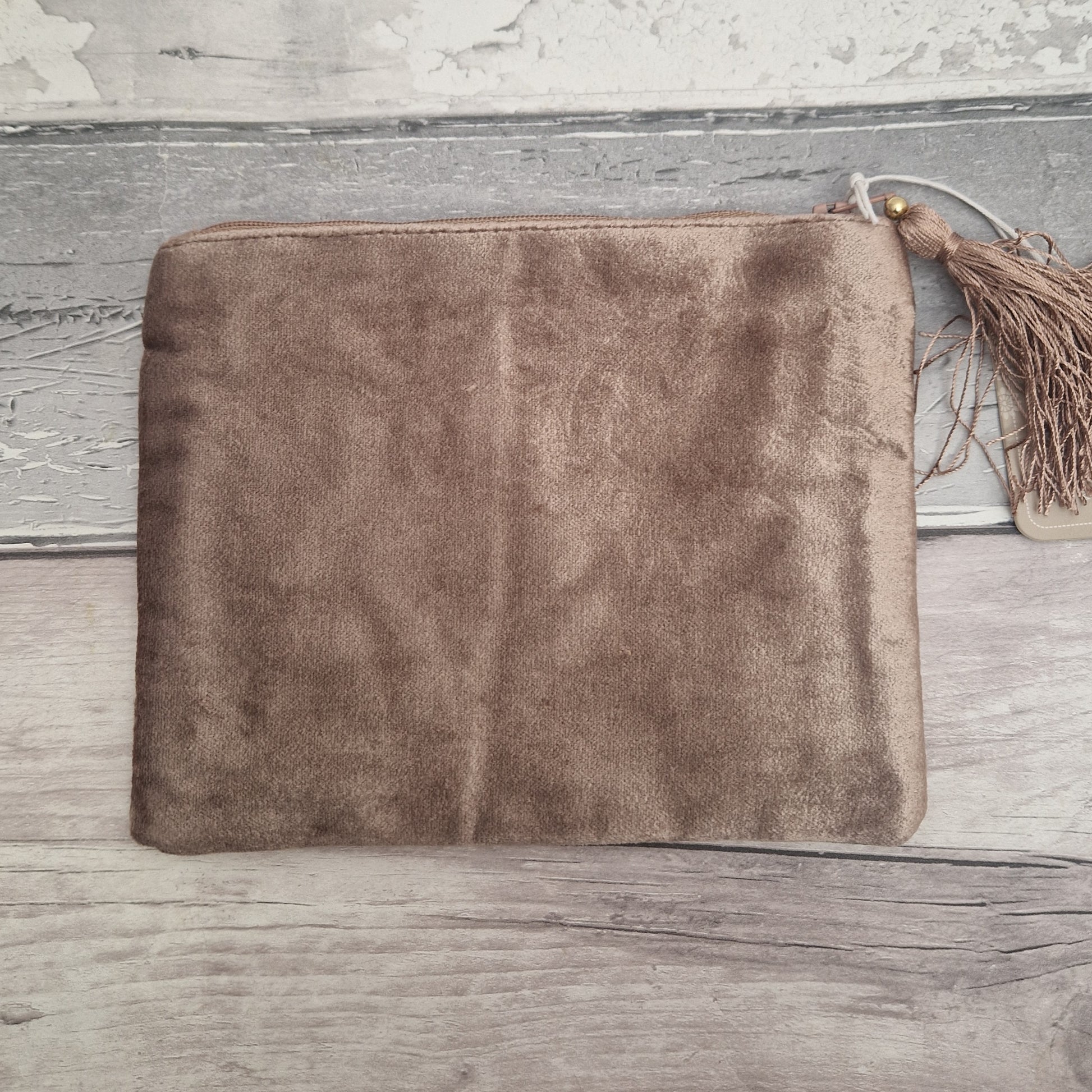 A brown velvet zippered pouch with a decorative tassel zipper pull, displayed flat on a rustic, weathered gray wood background.