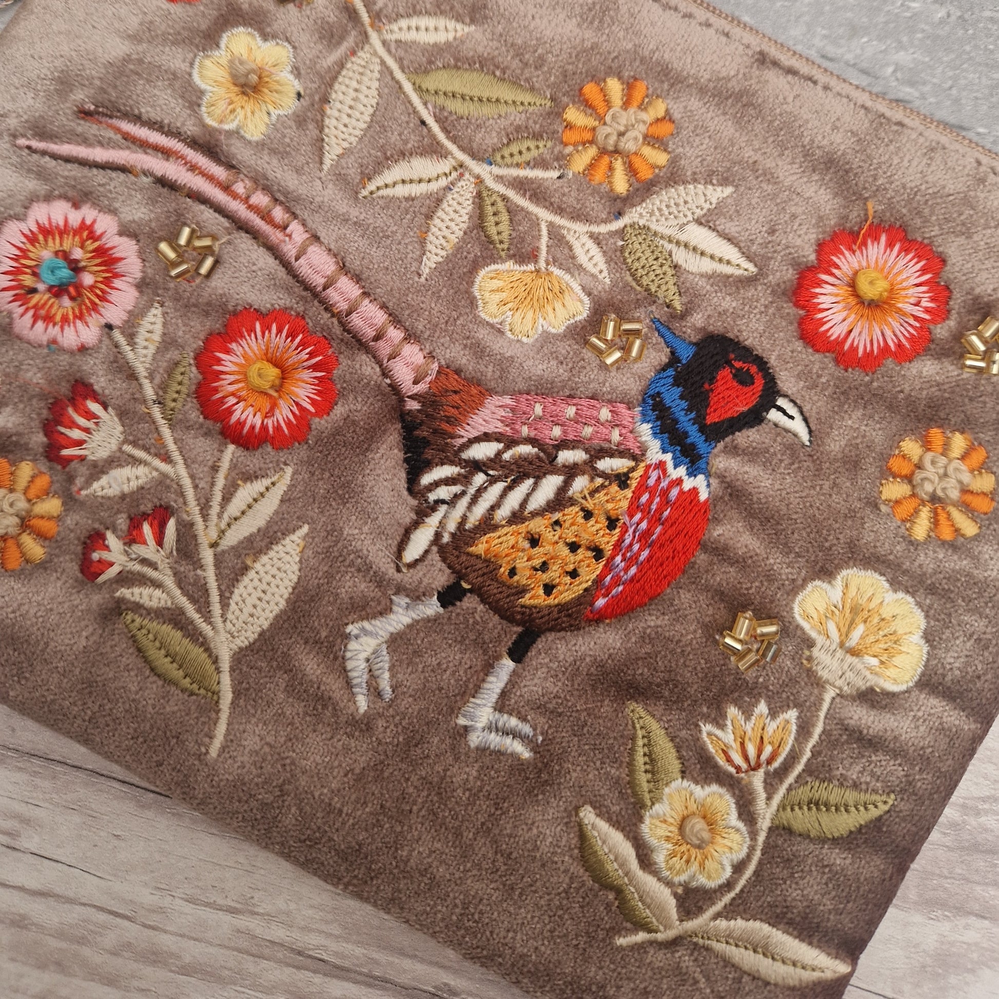 A close-up of a brown embroidered textile featuring a pheasant bird and floral motifs in red, yellow, cream, and orange on a textured fabric surface.