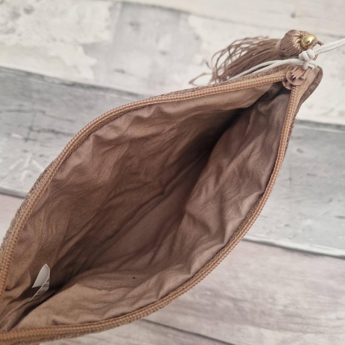 A close-up of an open brown zippered pouch or clutch, showing the interior lining and a decorative tassel with a gold bead on the zipper pull, set against a light gray wood plank background.