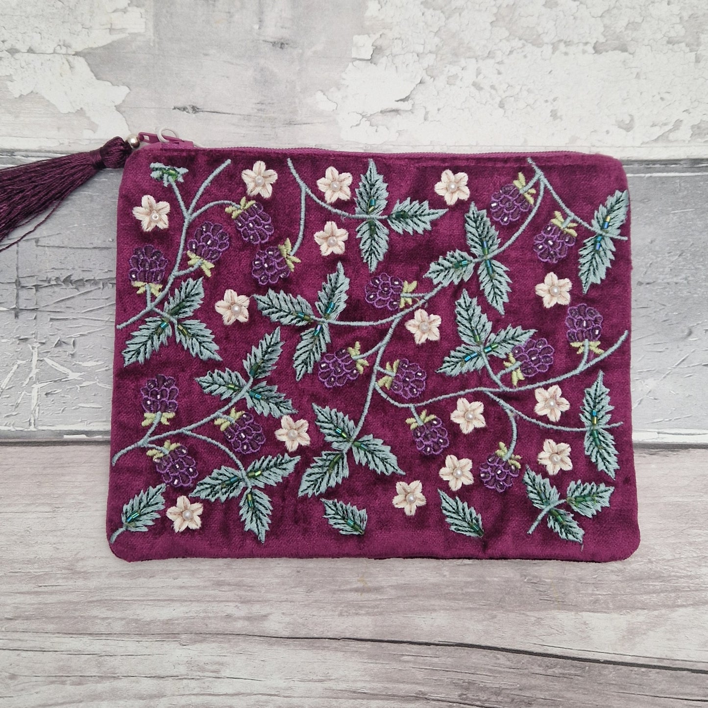 A rectangular velvet pouch in deep burgundy with embroidered raspberry and leaf patterns in green, purple, and cream, featuring a matching tassel zipper pull, laid flat on a rustic gray wood surface.