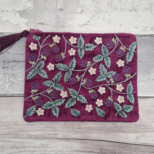 A rectangular velvet pouch in deep burgundy with embroidered raspberry and leaf patterns in green, purple, and cream, featuring a matching tassel zipper pull, laid flat on a rustic gray wood surface.