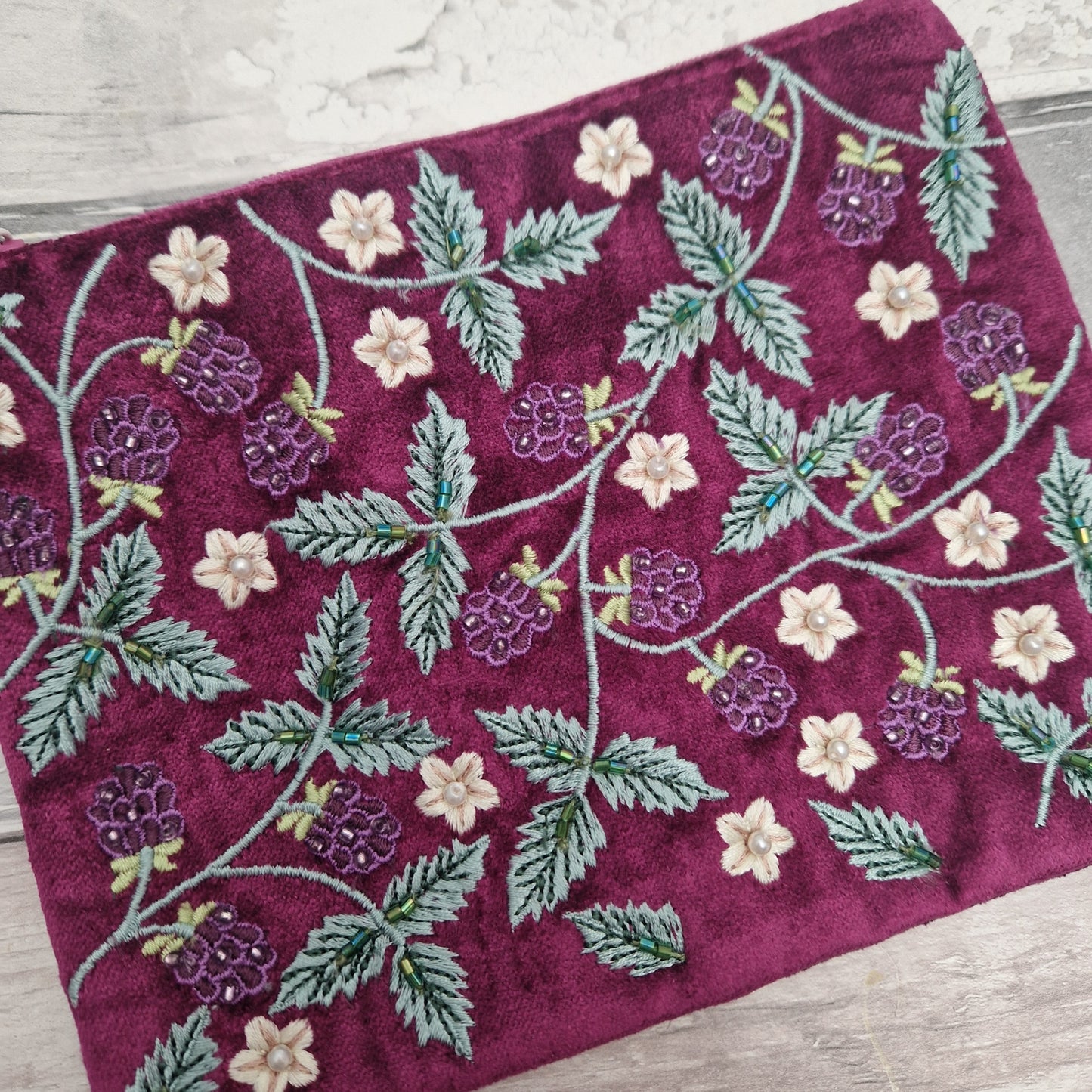 A close-up of a deep magenta velvet fabric featuring embroidered raspberry and floral vine patterns in green, purple, and cream, laid flat on a light wood surface.