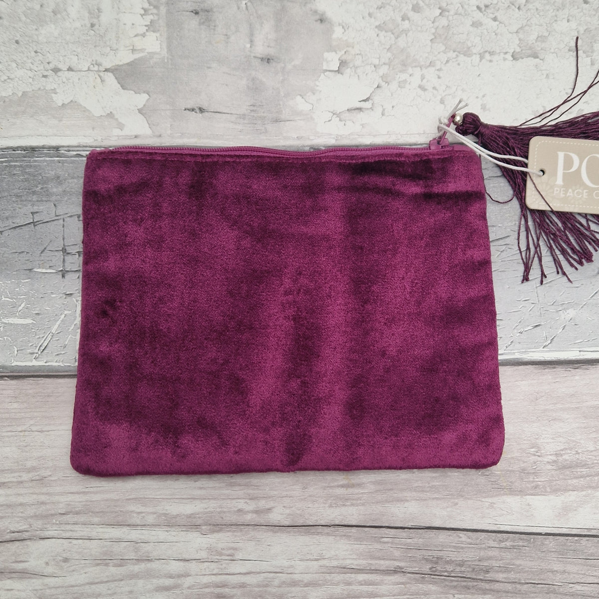 A deep magenta velvet pouch with a matching tassel and a paper tag, displayed on a rustic, light gray wood and plaster background.