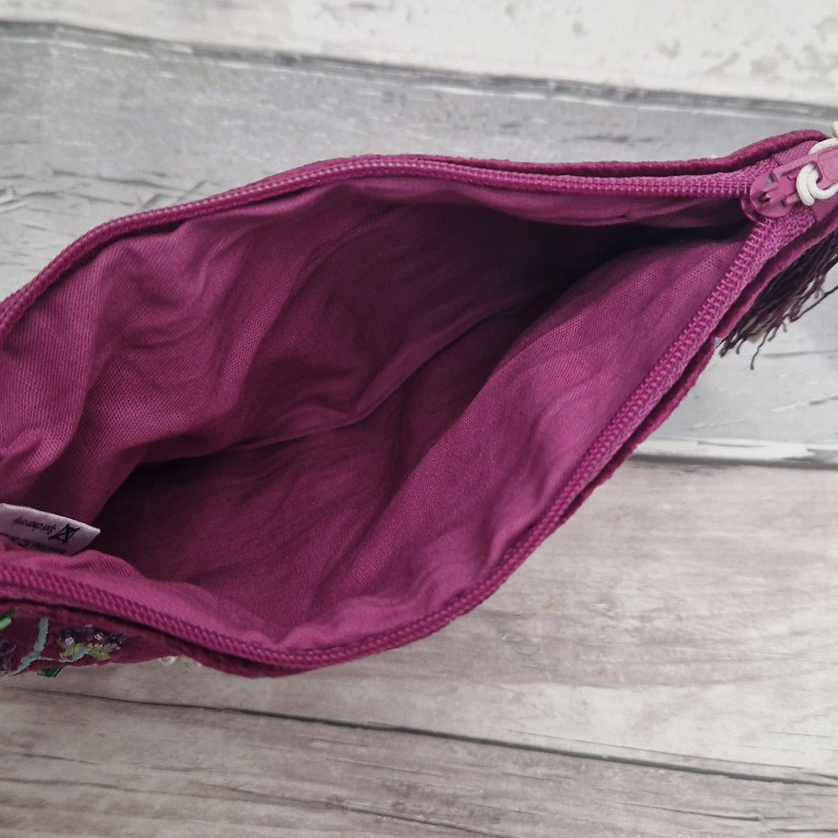 A close-up of an open burgundy fabric pouch or clutch, showing the interior lining and zipper, placed on a light gray wood surface.