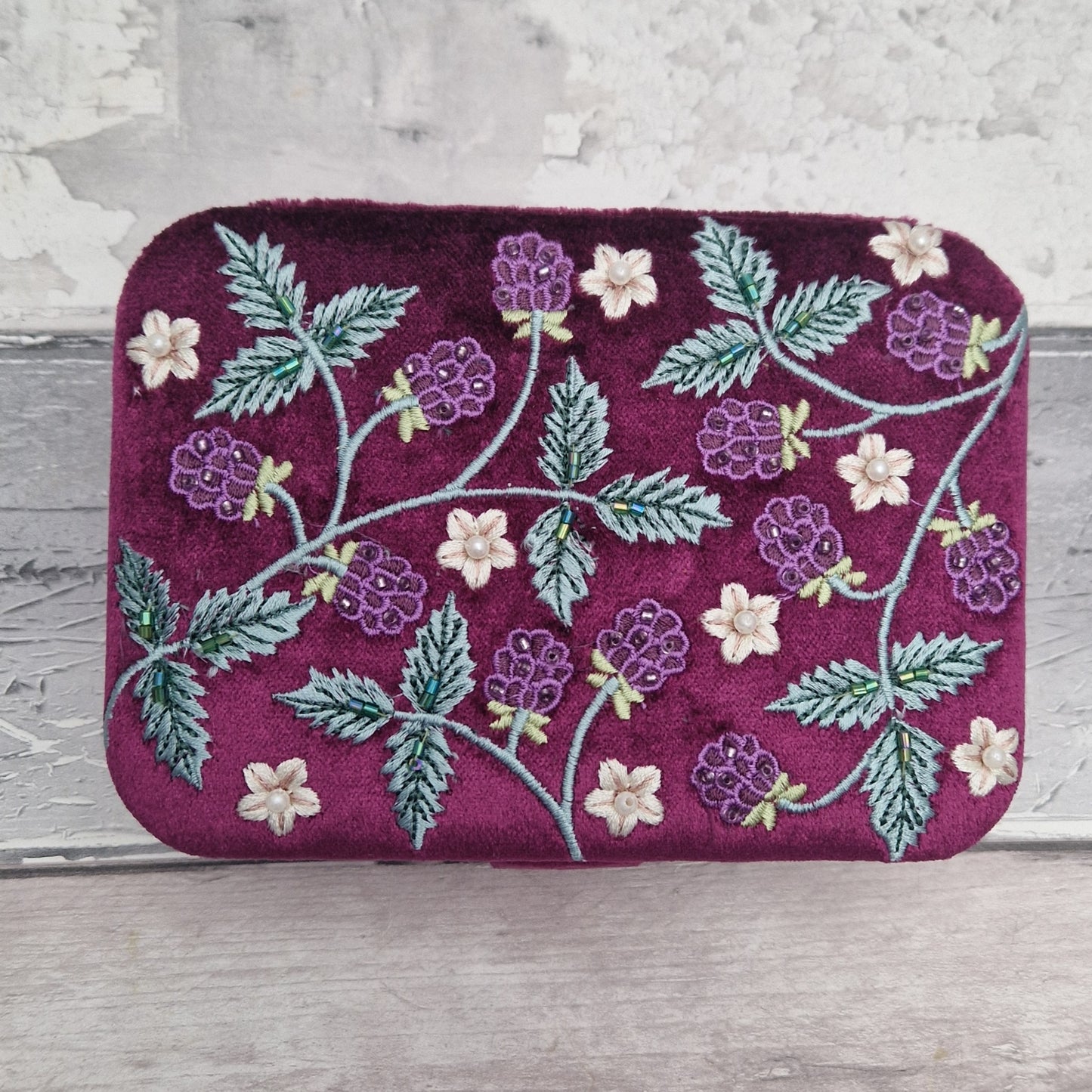 A rectangular velvet jewellery box in deep magenta-purple, decorated with embroidered purple blackberries, green leaves, and small cream flowers, set against a rustic light gray wooden background.