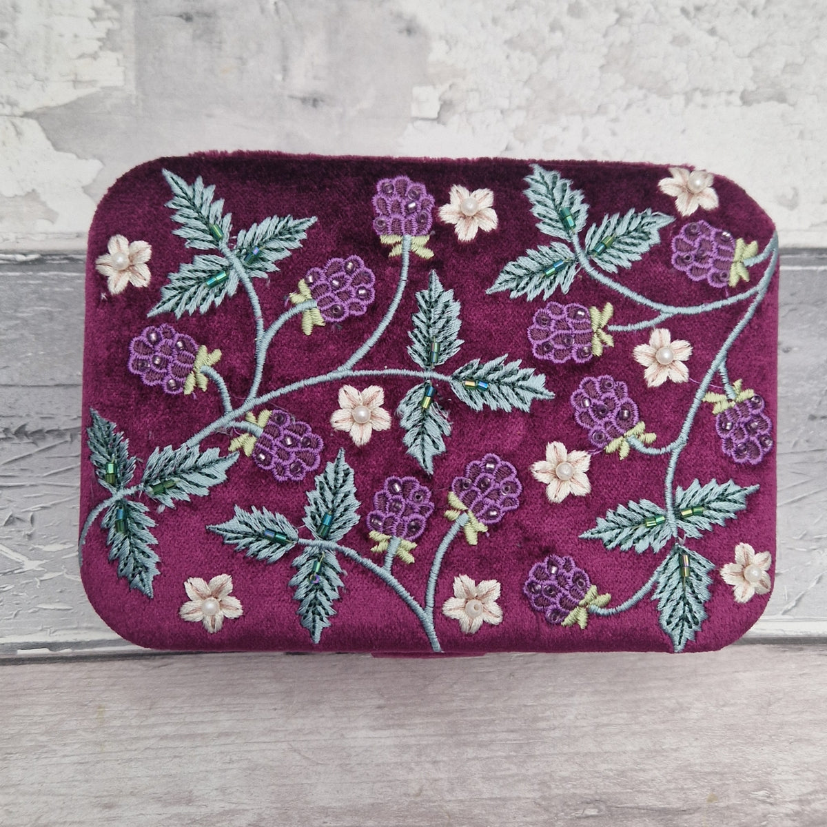 A rectangular velvet jewellery box in deep magenta-purple, decorated with embroidered purple blackberries, green leaves, and small cream flowers, set against a rustic light gray wooden background.