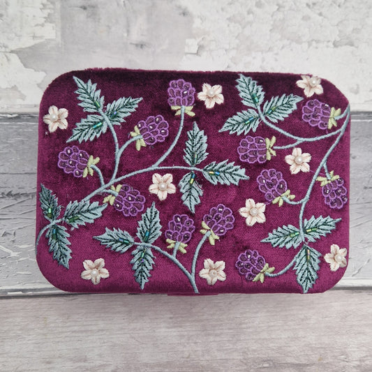 A rectangular velvet jewellery box in deep magenta-purple, decorated with embroidered purple blackberries, green leaves, and small cream flowers, set against a rustic light gray wooden background.
