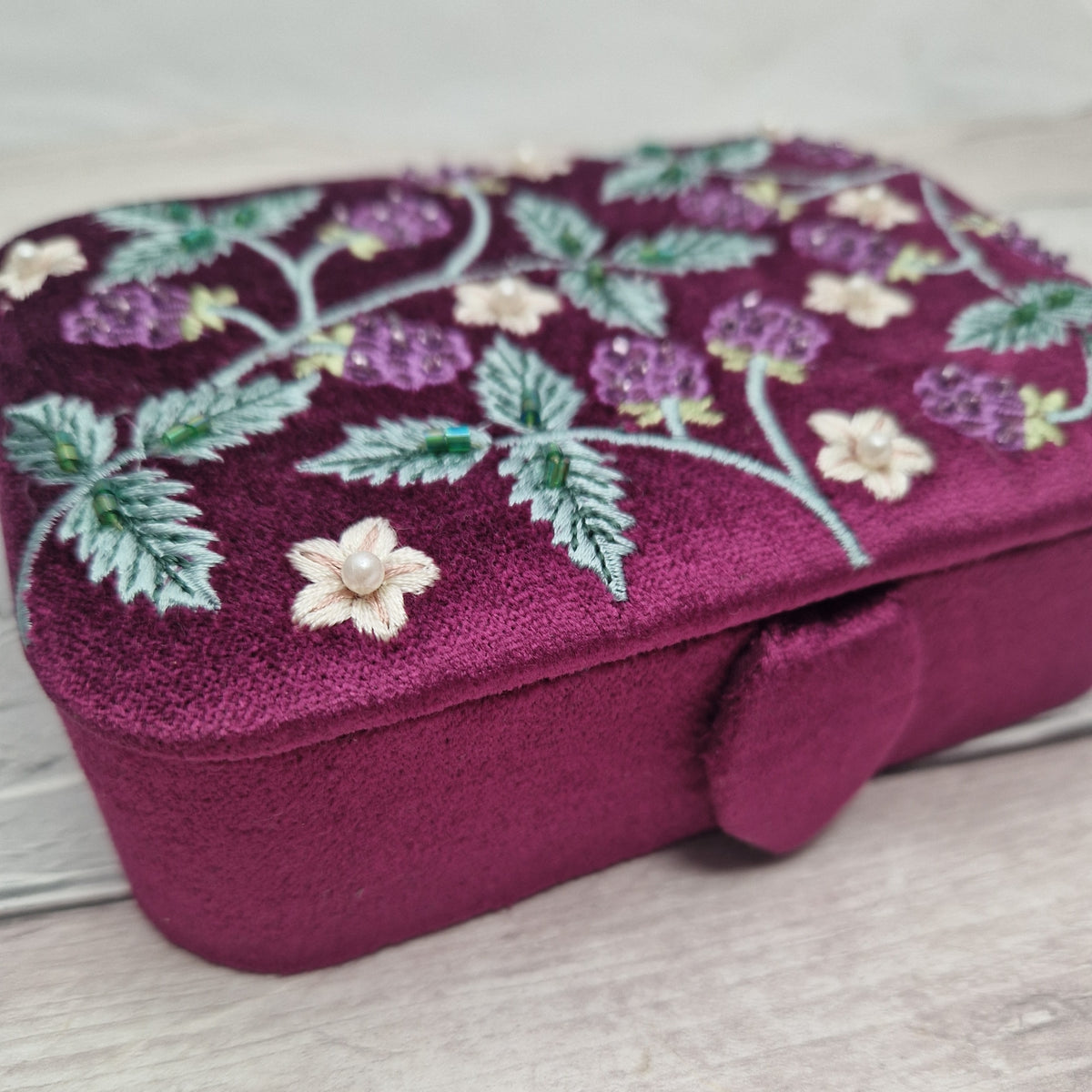 A rectangular velvet jewelry box in deep magenta, decorated with embroidered purple and cream flowers and green leaves on the lid, resting on a light wood surface.