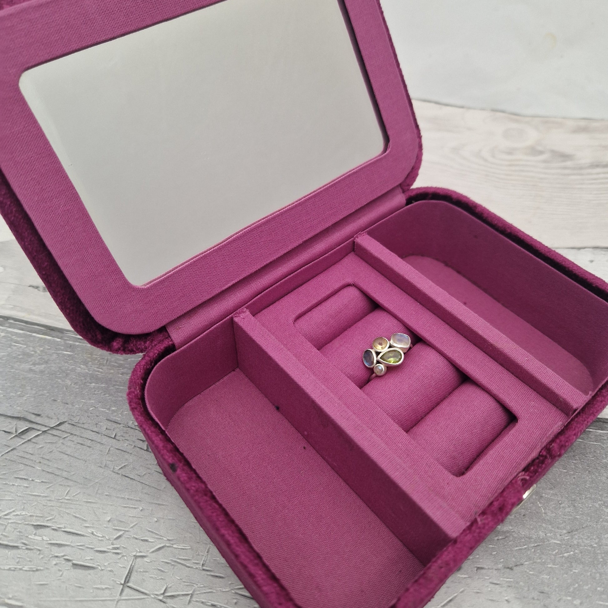 A magenta velvet jewellery box with an interior mirror, divided compartments, and a ring holder containing a gold ring, set on a light gray textured surface.