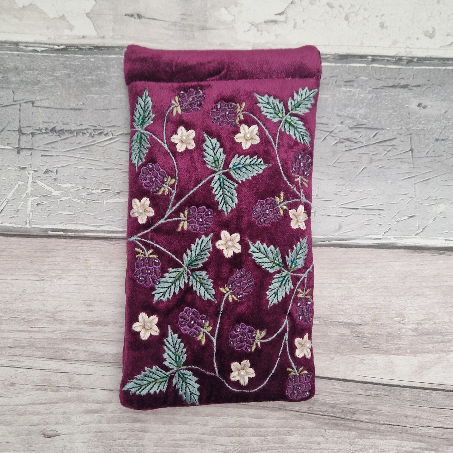 A deep burgundy velvet glasses case or sleeve featuring embroidered berry and leaf patterns in green, purple, and cream, displayed on a light gray wooden surface.