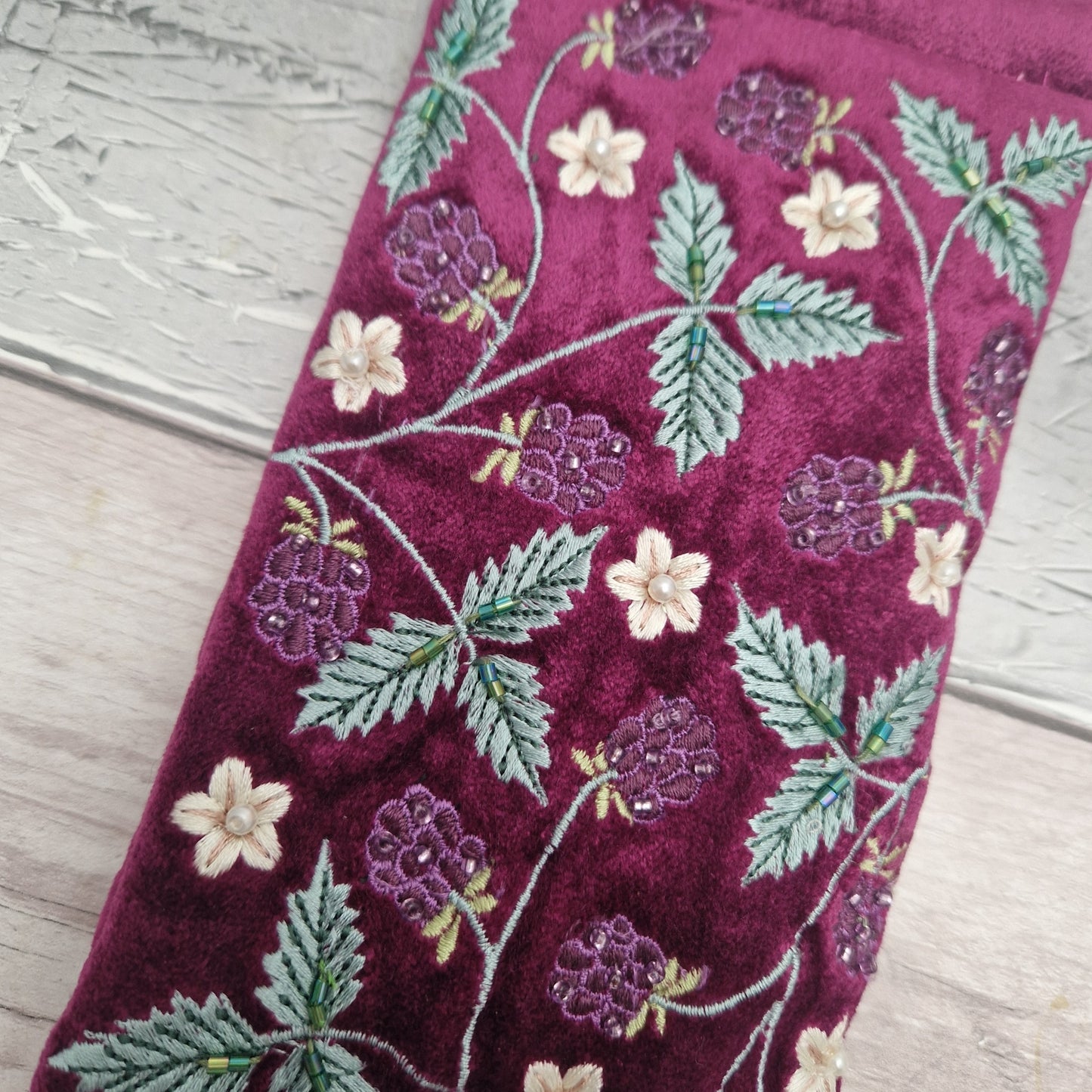 A close-up of a deep burgundy velvet glasses case featuring embroidered raspberry and flower motifs in purple, green, and cream, laid flat on a light wood surface.