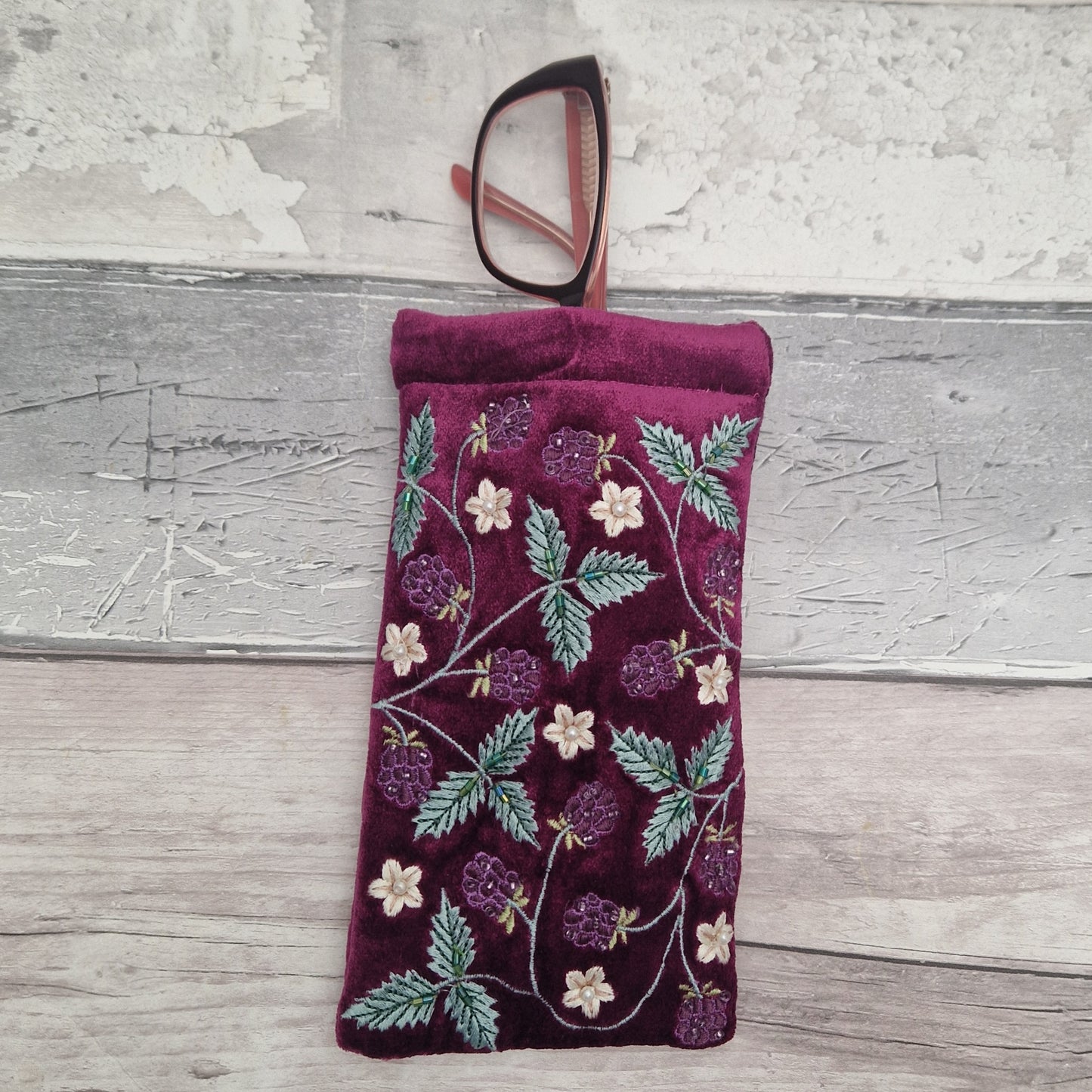 A burgundy velvet eyeglasses pouch with embroidered green leaves and purple berries, shown with a pair of eyeglasses resting inside the top opening, set against a rustic gray wood background.