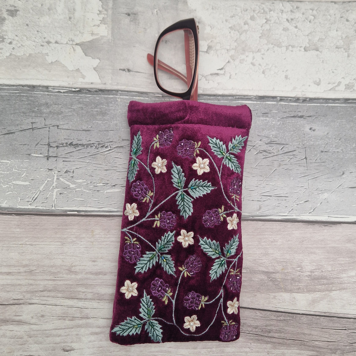 A burgundy velvet eyeglasses pouch with embroidered green leaves and purple berries, shown with a pair of eyeglasses resting inside the top opening, set against a rustic gray wood background.