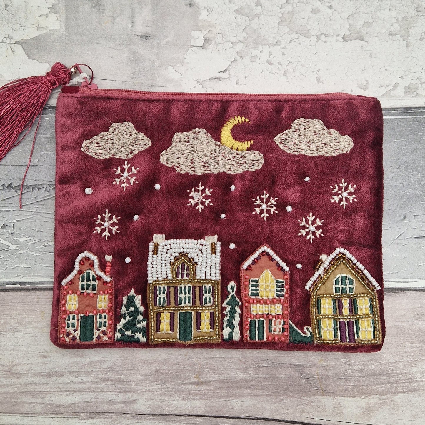 A burgundy velvet zippered pouch with a tassel, decorated with an embroidered winter village scene featuring houses, snowflakes, clouds, a crescent moon, and pine trees, displayed on a rustic wooden surface.