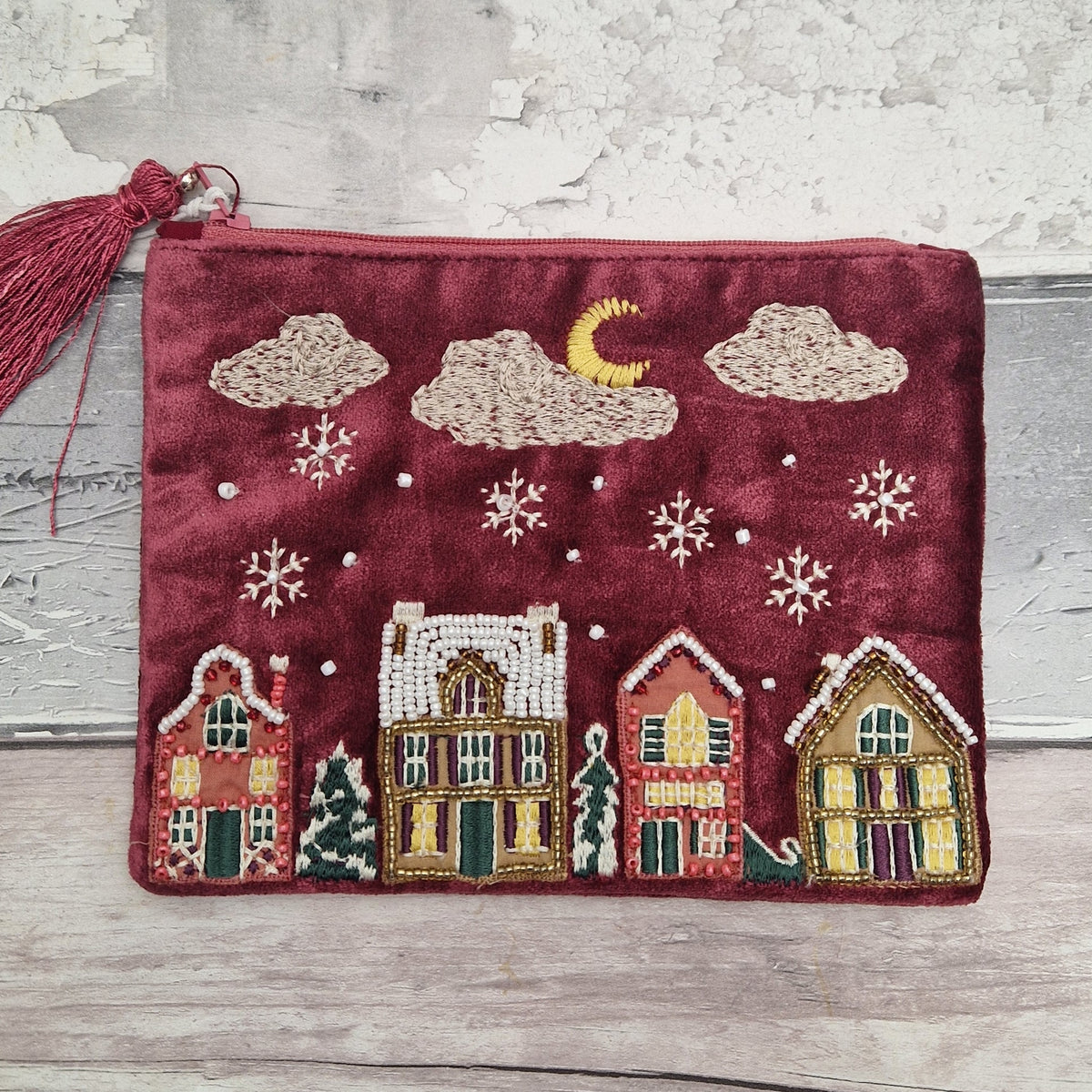 A burgundy velvet zippered pouch with a tassel, decorated with an embroidered winter village scene featuring houses, snowflakes, clouds, a crescent moon, and pine trees, displayed on a rustic wooden surface.