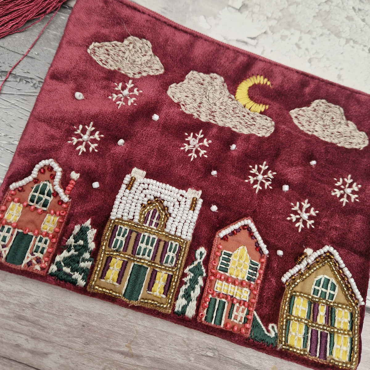 A close-up of a red embroidered textile featuring four decorated houses, snowflakes, clouds, a crescent moon, and small trees, laid on a light wood surface.
