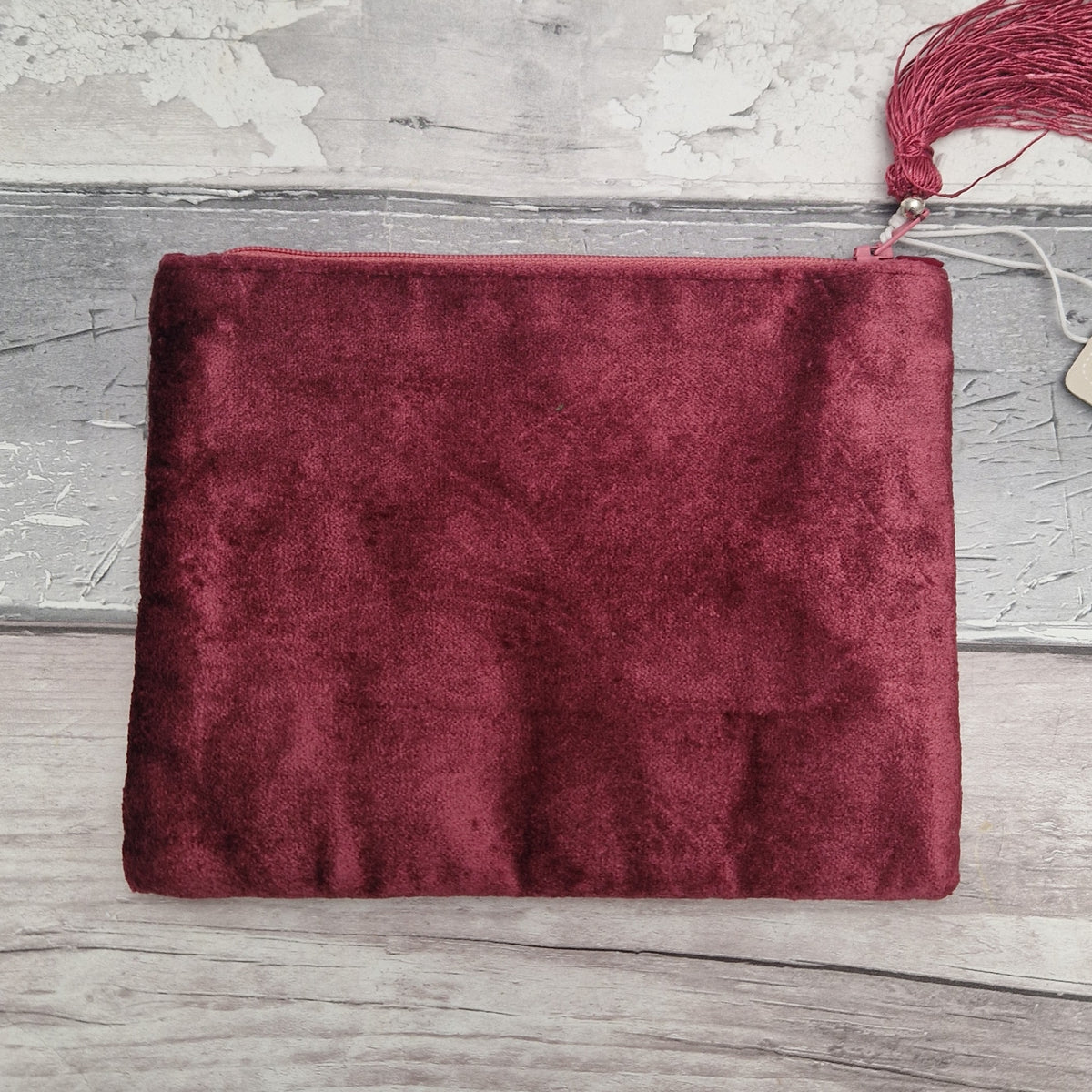 A rectangular burgundy velvet pouch or clutch with a matching tassel zipper pull, laid flat on a rustic gray wooden surface.