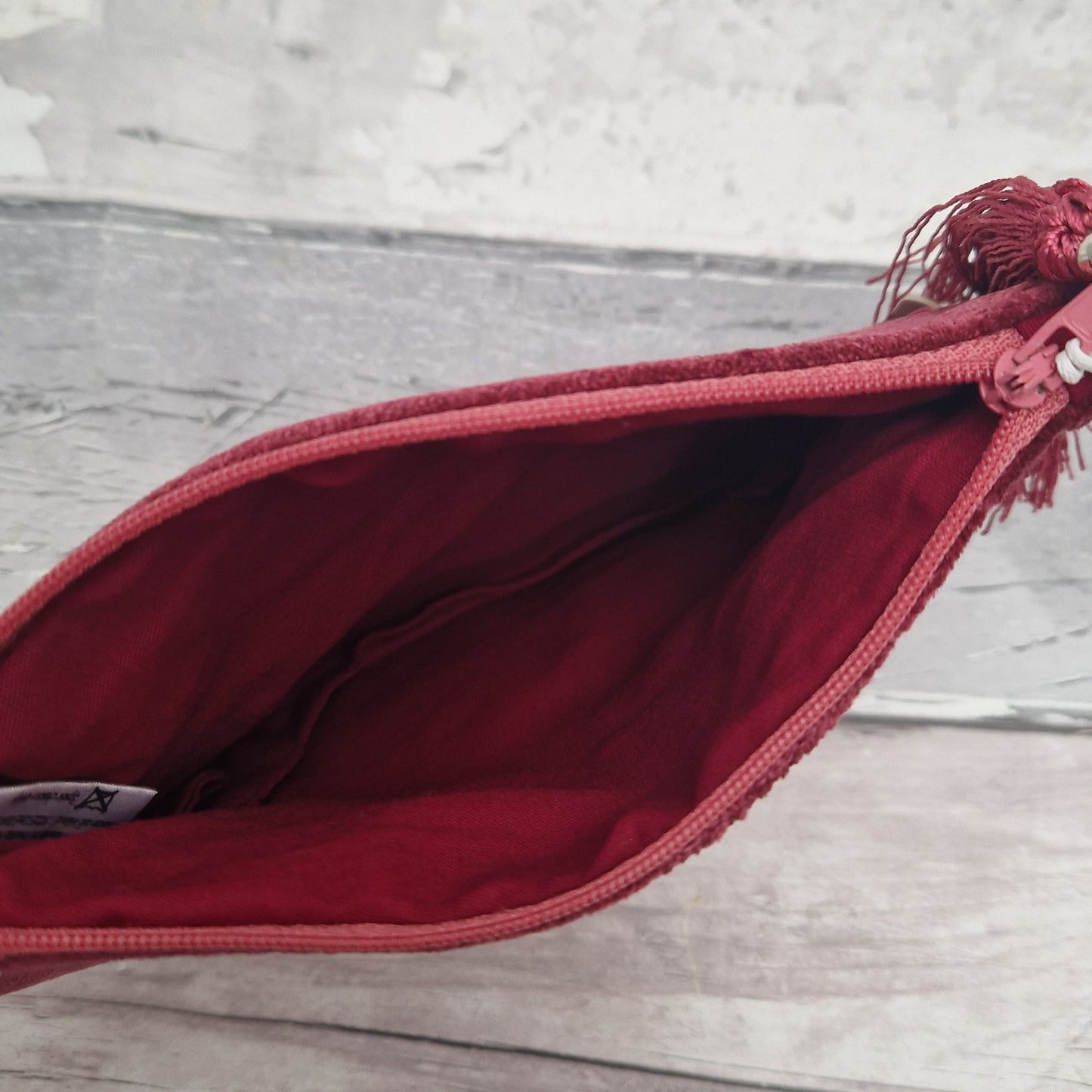 A close-up of an open burgundy zippered pouch or clutch, showing the interior lining and zipper tape, with a matching tassel detail at the top right, set against a light gray wood-textured background.