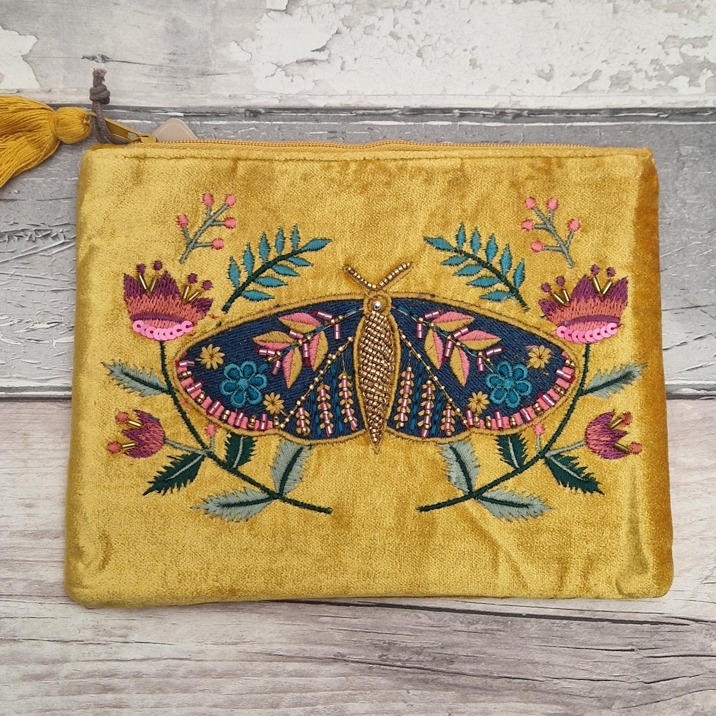 A yellow velvet zippered pouch featuring a large embroidered moth and floral design in pink, blue, green, and gold, resting on a rustic gray wooden surface.
