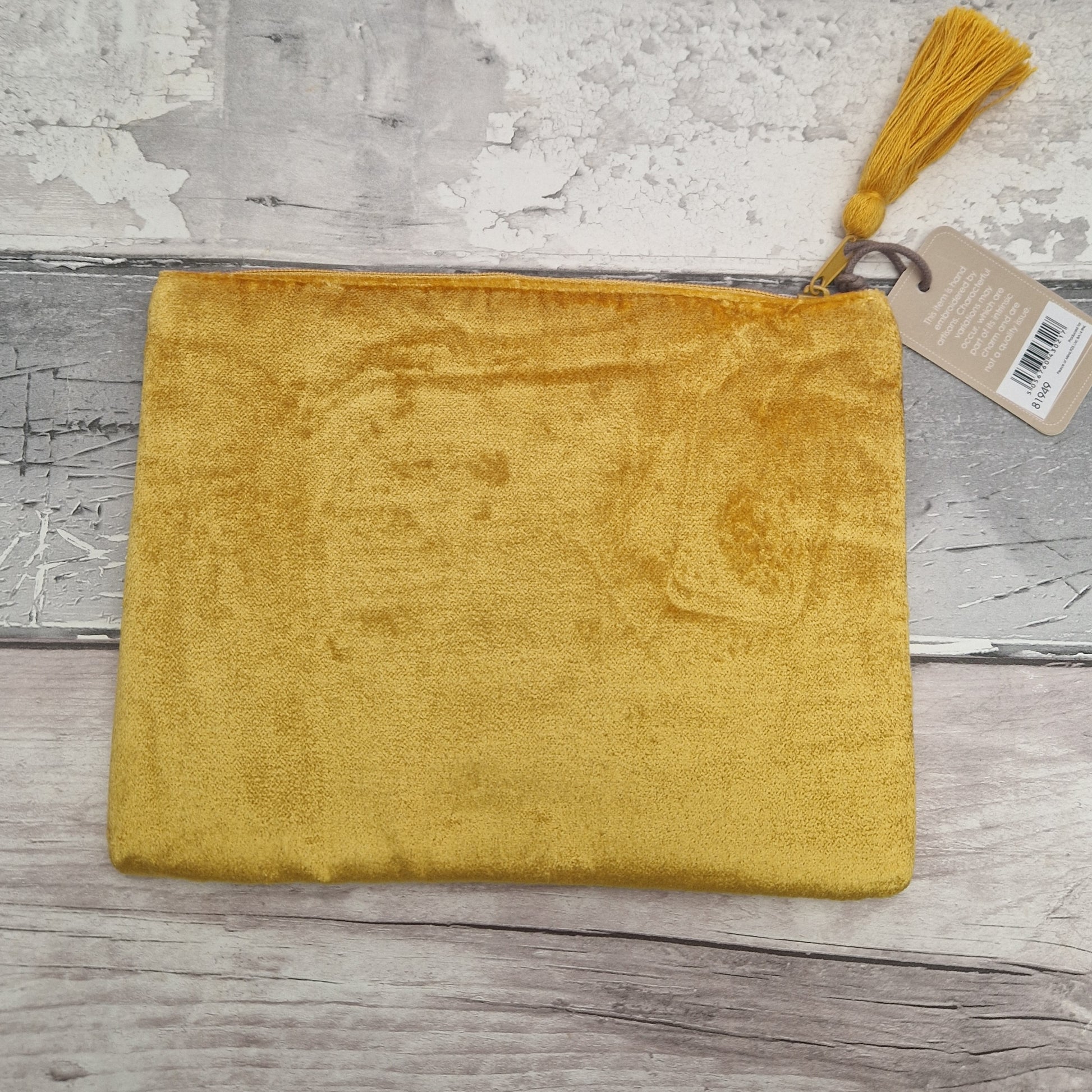 A mustard yellow velvet pouch with a top zipper, a matching tassel, and a paper tag, displayed on a rustic gray wood background.