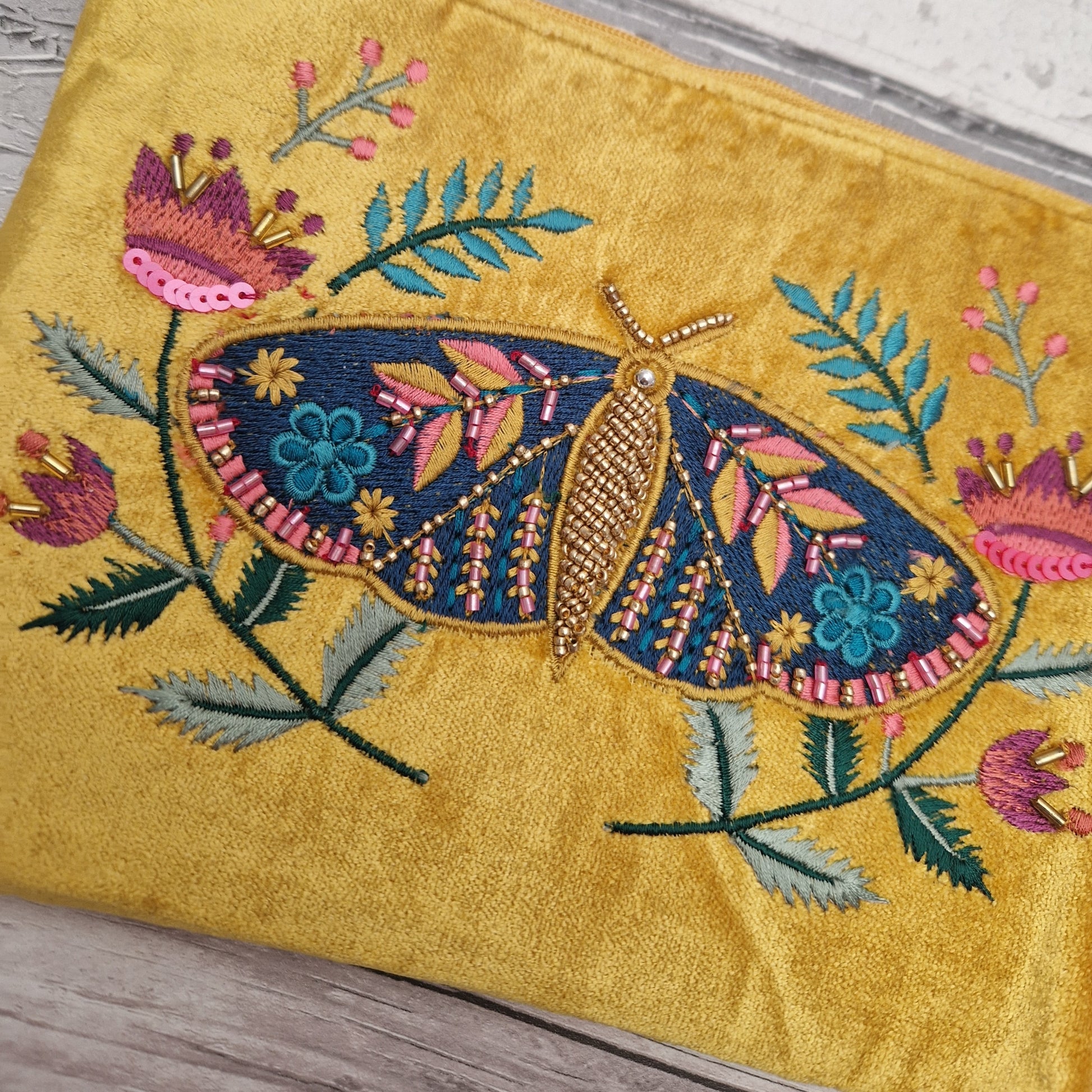 A close-up of a mustard yellow velvet pouch featuring a large, colorful embroidered moth and floral motifs on the front.