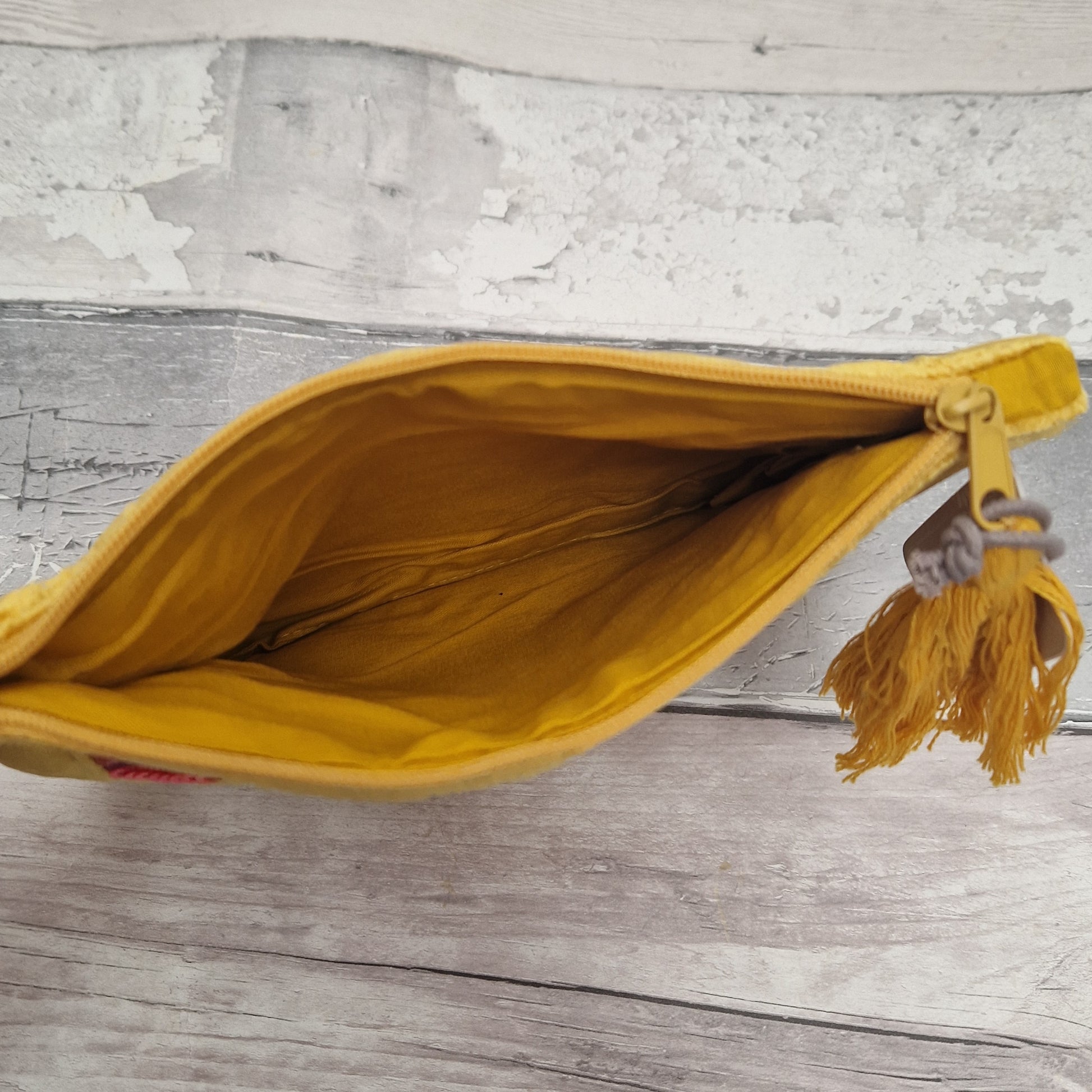 A mustard yellow fabric pouch or clutch shown open, revealing a matching yellow interior and a tassel zipper pull, placed on a light gray wood surface.