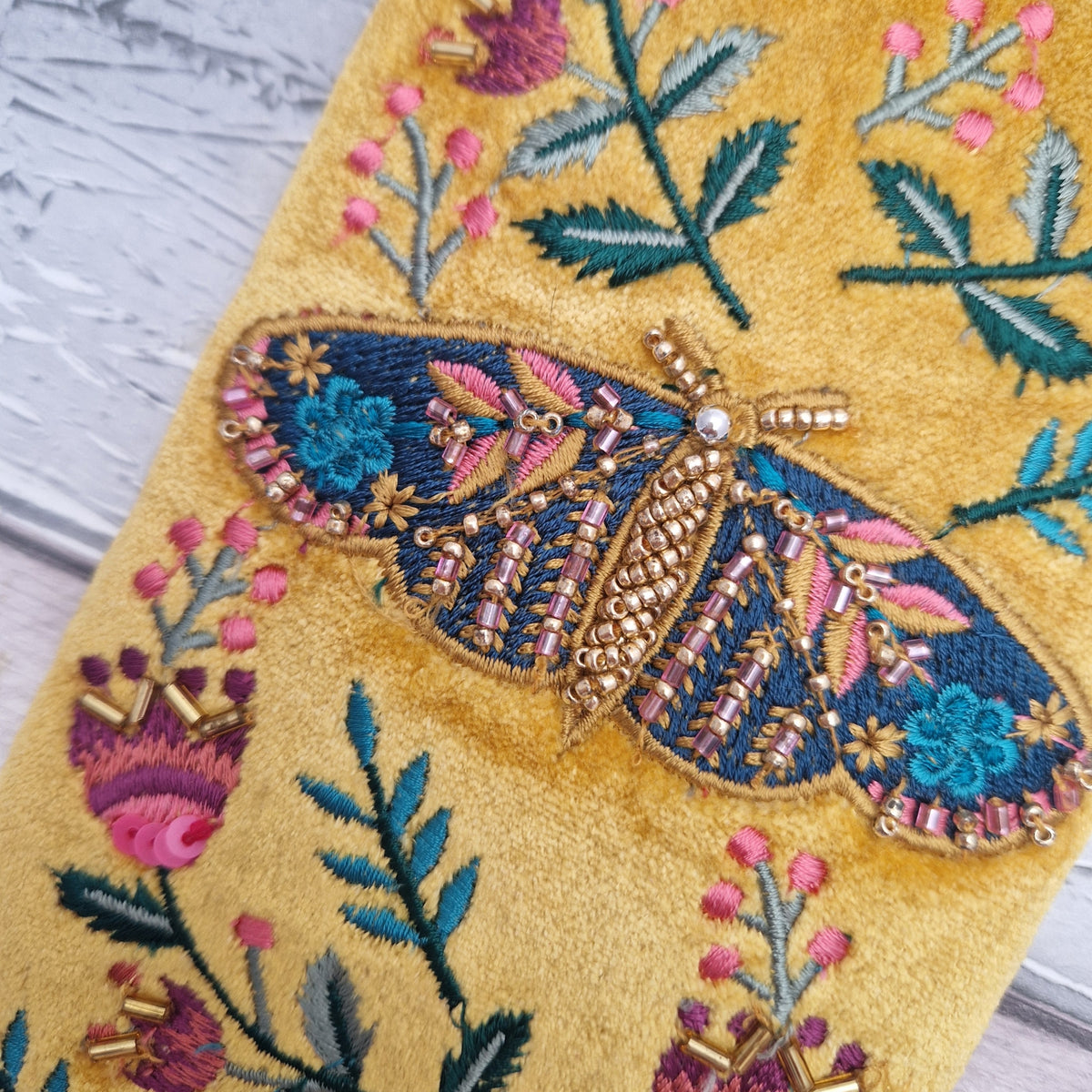 A close-up of a yellow embroidered textile featuring a large decorative moth with blue, pink, and gold embroidered wings, surrounded by floral motifs on a textured fabric.