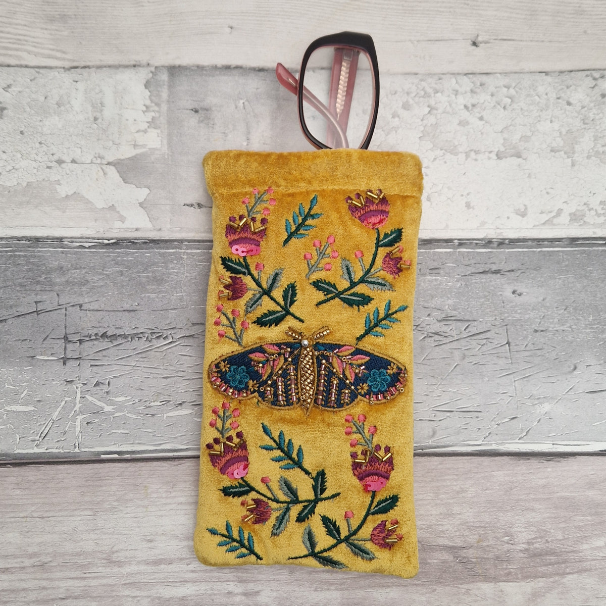 A yellow velvet eyeglasses pouch with colorful floral and butterfly embroidery, shown with a pair of eyeglasses resting inside, set against a rustic white-washed wood background.