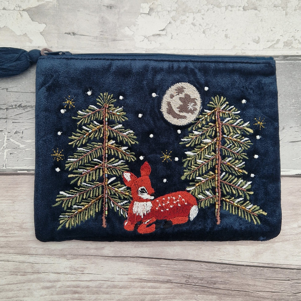 A navy blue velvet zippered pouch featuring a festive embroidered design of a red fawn, pine trees, stars, and a moon, displayed on a rustic light wood background.