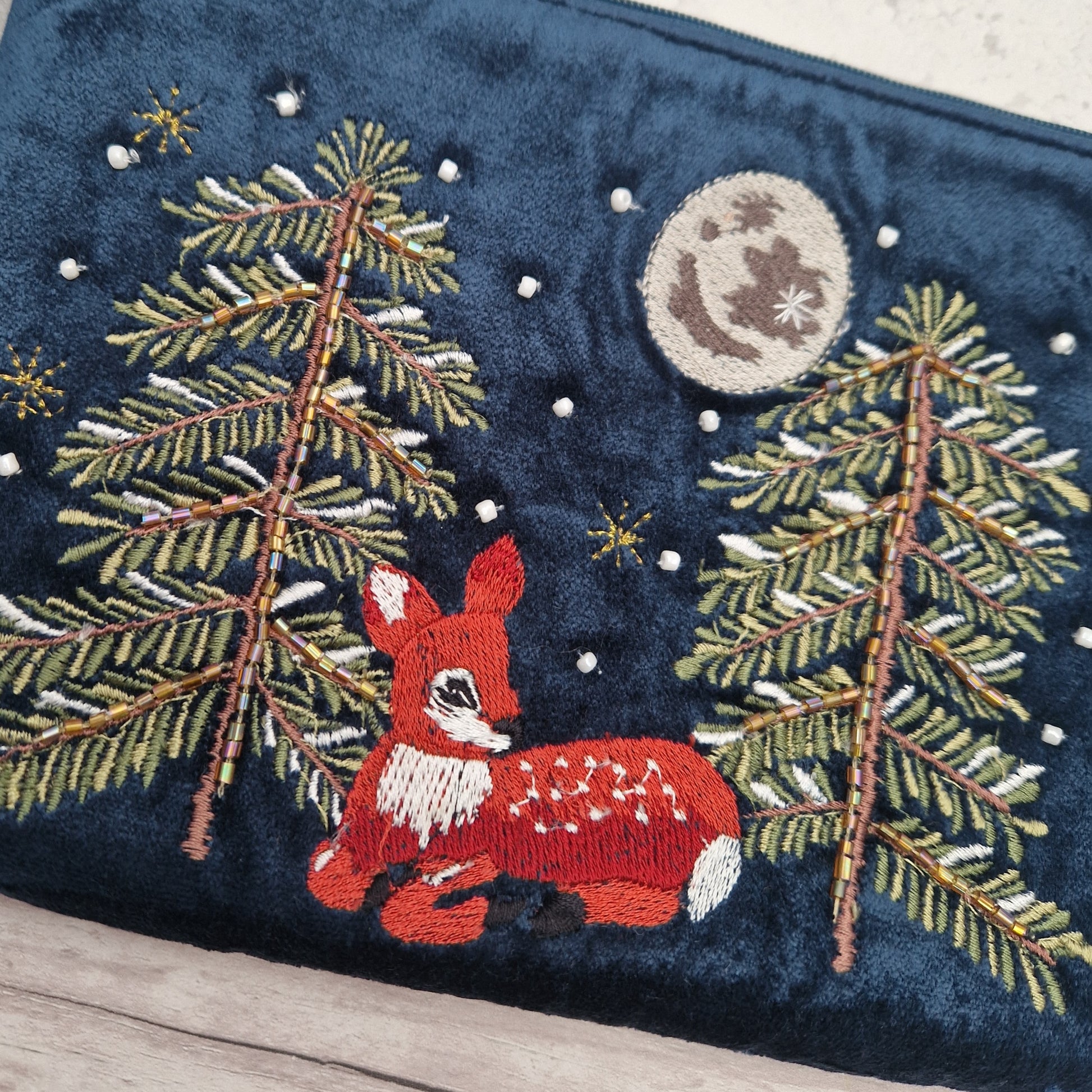 A close-up of a navy blue embroidered fabric item, likely a clutch bag or pouch, featuring a festive woodland design with a red fawn, pine trees, a moon, and snowflakes.