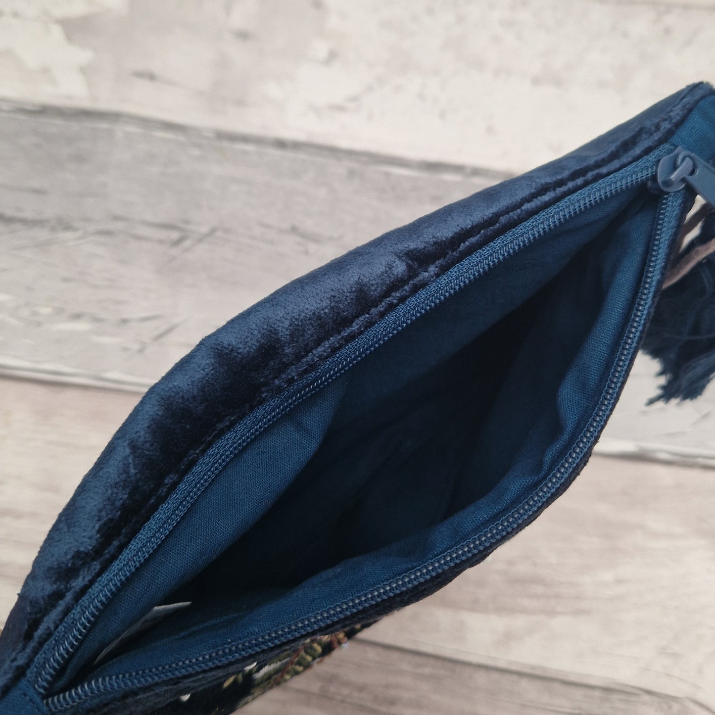 A close-up of a dark navy blue velvet pouch or clutch shown open, revealing its matching fabric zipper and interior lining, set against a light gray wood plank background.