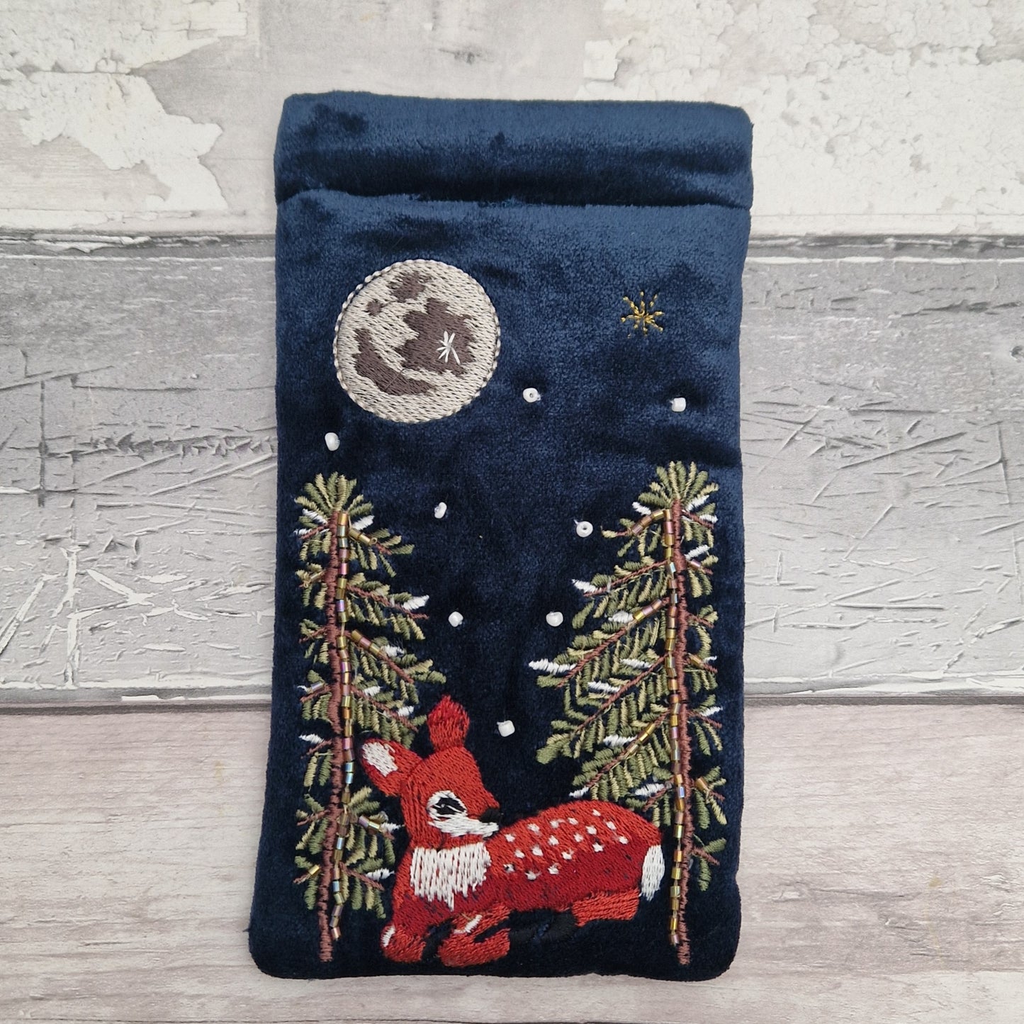 A dark navy blue velvet pouch or sleeve featuring a detailed embroidered Christmas night scene with a red and white fawn, pine trees, a full moon, and stars, displayed on a light gray wooden surface.