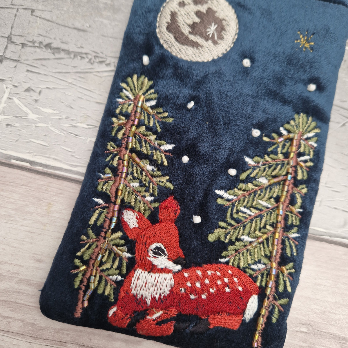 A close-up of a dark navy blue embroidered fabric item, likely a stocking or decorative textile, featuring a red and white spotted deer, green pine trees, and a cream-colored moon with a star on a light wood background.